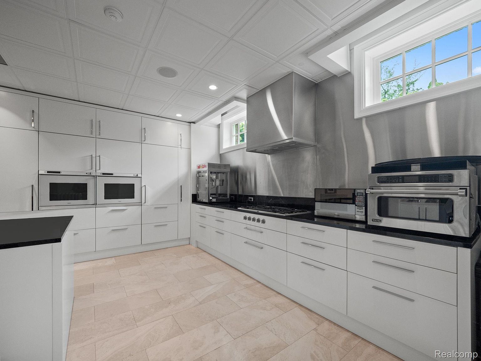 This is a well-lit kitchen featuring white cabinetry, black countertops, and stainless steel appliances. The kitchen has a modern design with a tiled ceiling and floor. Natural light enters through a window above the countertop.