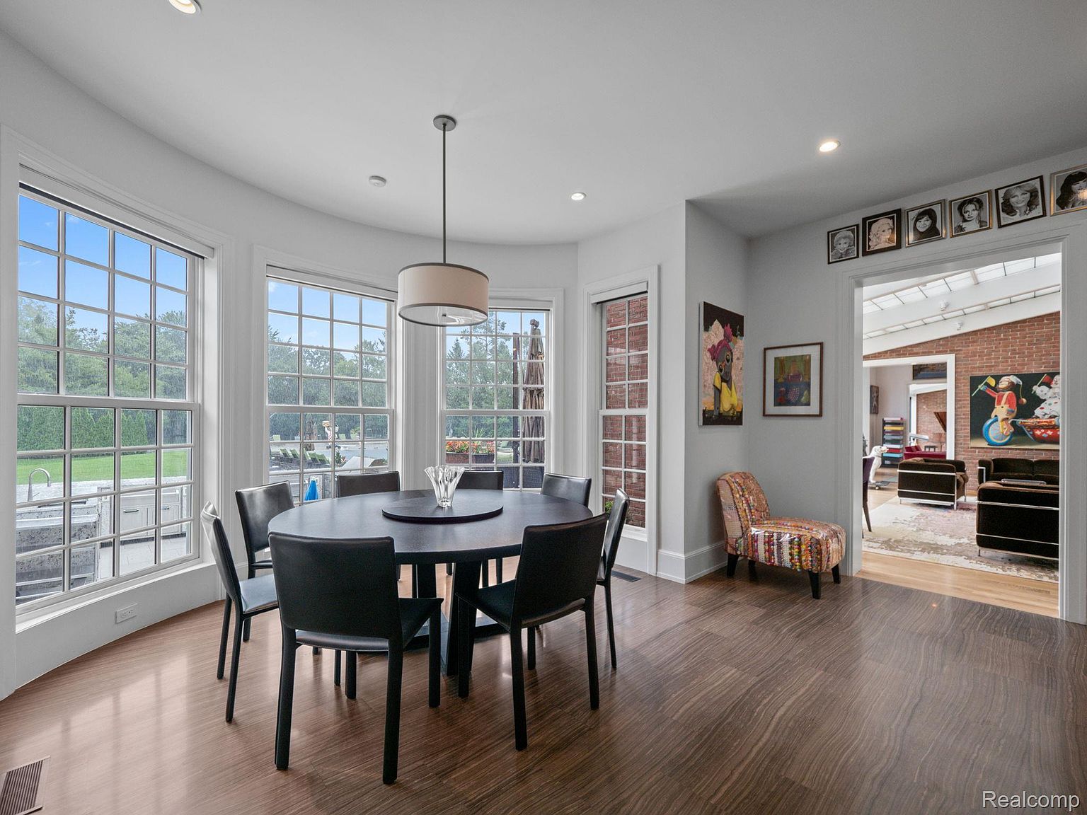 This is an interior shot of a dining room featuring a round, dark-colored dining table surrounded by black chairs. Large windows offer ample natural light and views of the exterior. The room has hardwood floors and a modern chandelier hangs above the table, creating an inviting and stylish space for dining.