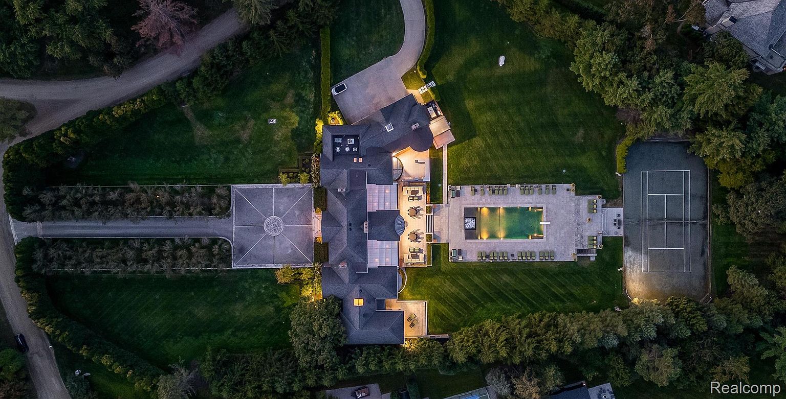 This aerial view showcases a sprawling estate with meticulously manicured grounds. The property features a large residence with a dark roof, a long driveway leading to a circular motor court, a swimming pool with surrounding lounge area, and a tennis court. Lush greenery and mature trees provide privacy and enhance the overall sense of luxury and seclusion.