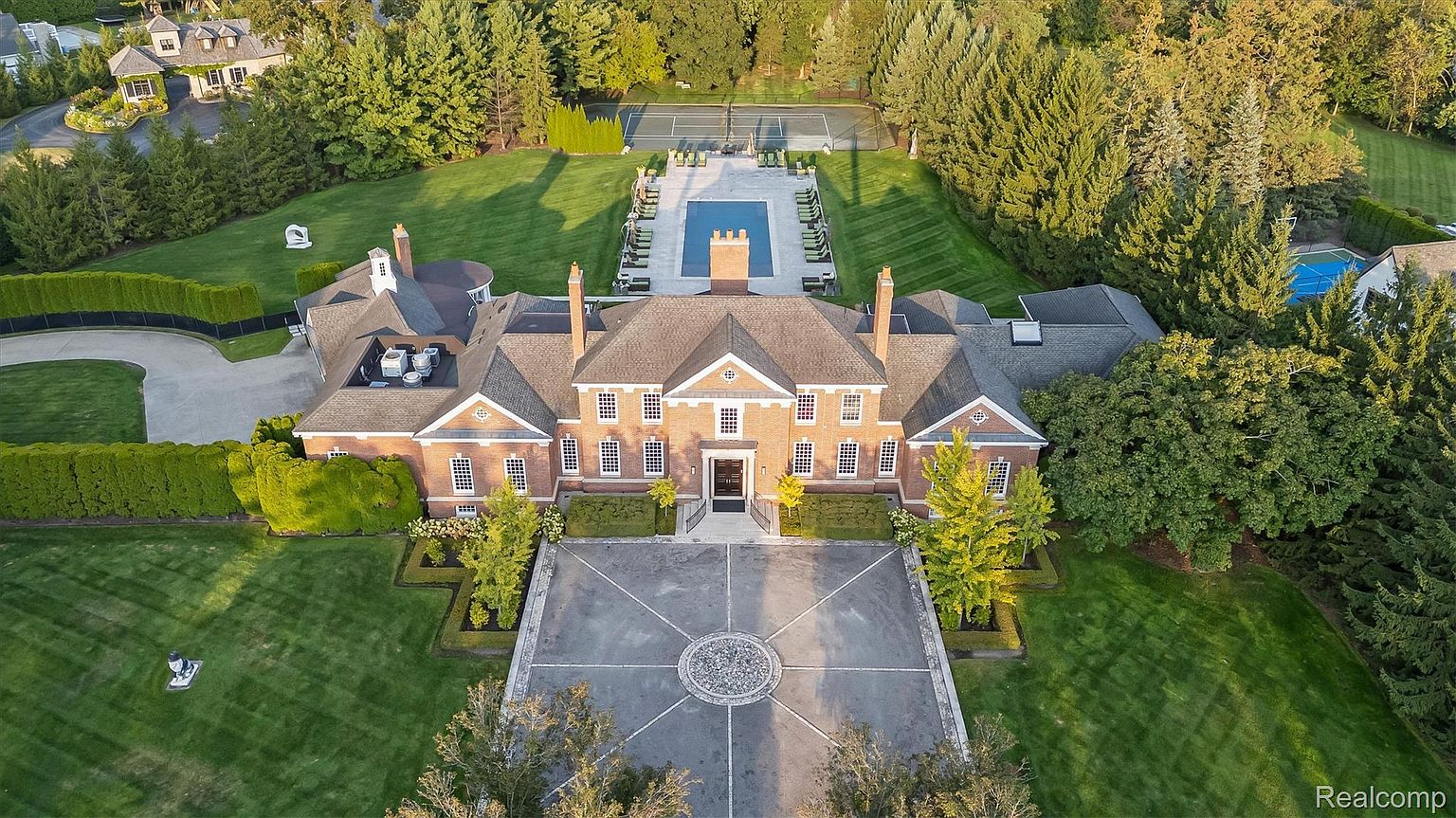 This aerial view showcases a grand brick mansion with a symmetrical design, complemented by meticulously manicured lawns and mature trees. A circular driveway leads to the entrance, while a pool and tennis court are visible in the rear, suggesting a luxurious and private estate. The overall impression is one of opulence and refined living.