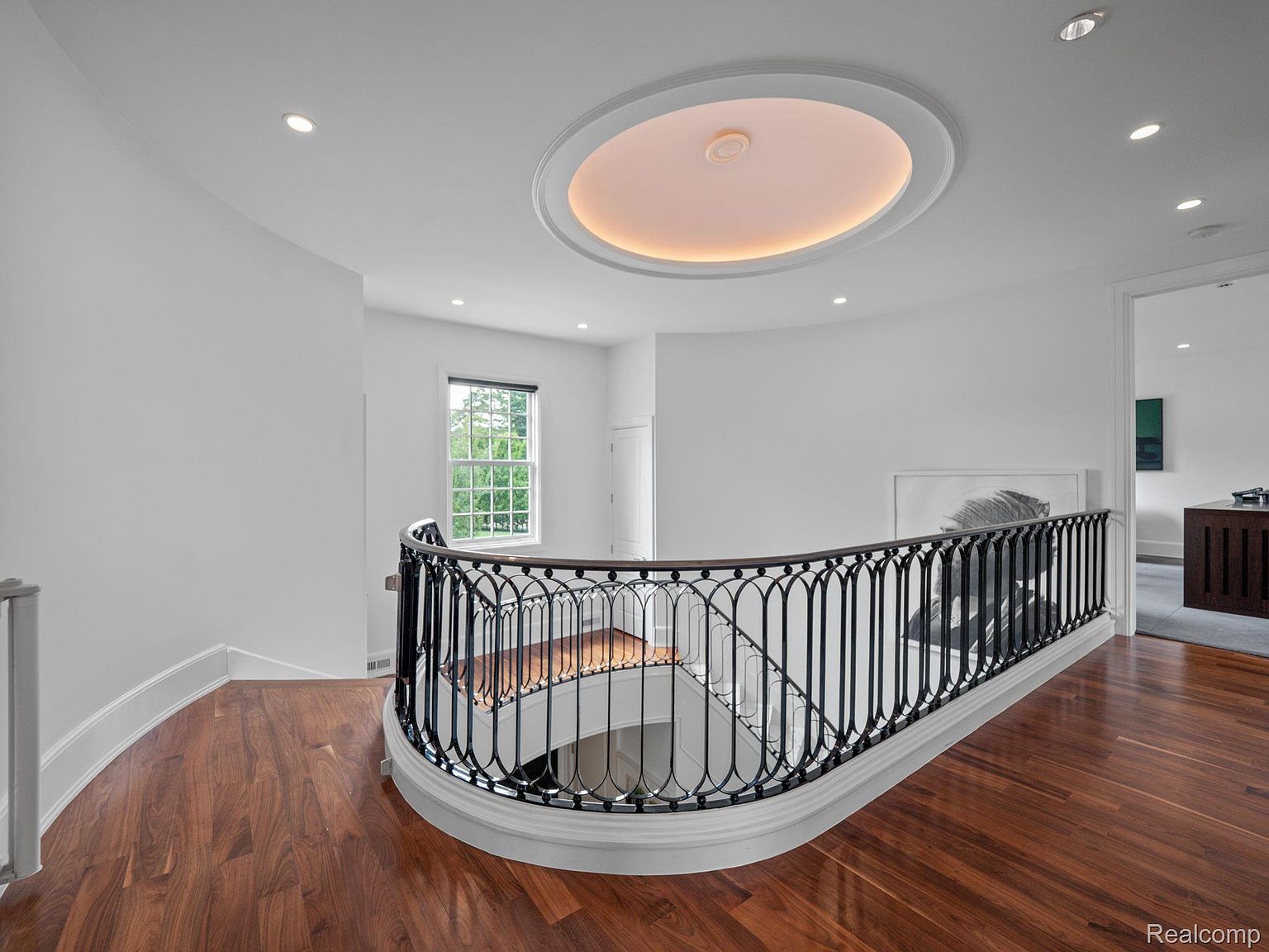 This is an interior shot of a hallway and staircase area. The space features hardwood flooring, white walls, and a black wrought iron railing. A unique oval ceiling light fixture adds a touch of elegance, and a window provides natural light. The overall impression is clean, bright, and sophisticated.
