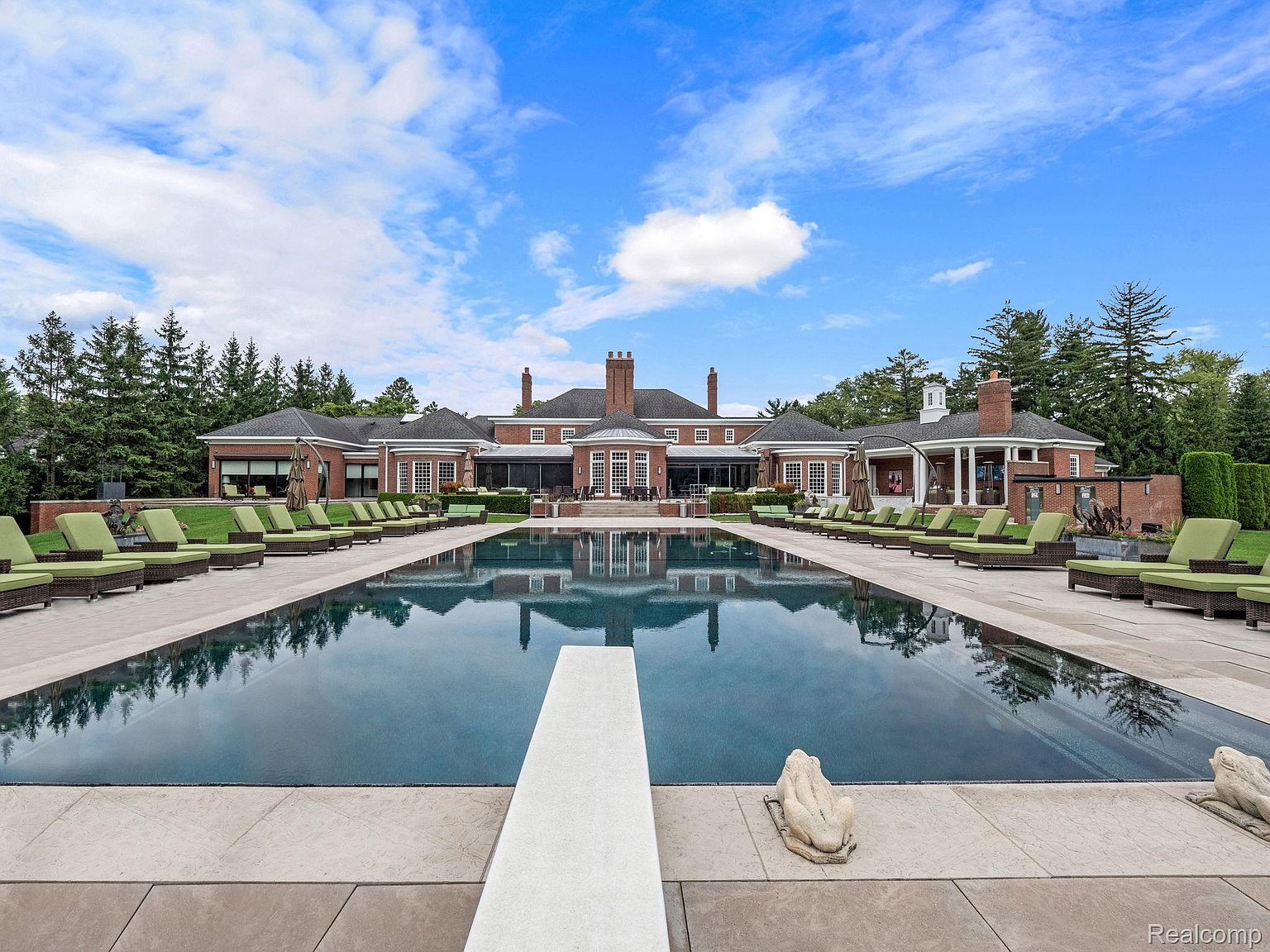 This image showcases a luxurious outdoor pool area with numerous lounge chairs arranged on both sides of the pool. The pool reflects the grand brick mansion in the background, which features multiple chimneys and a symmetrical design. A diving board is visible at the front of the pool, and decorative statues add an elegant touch to the poolside.