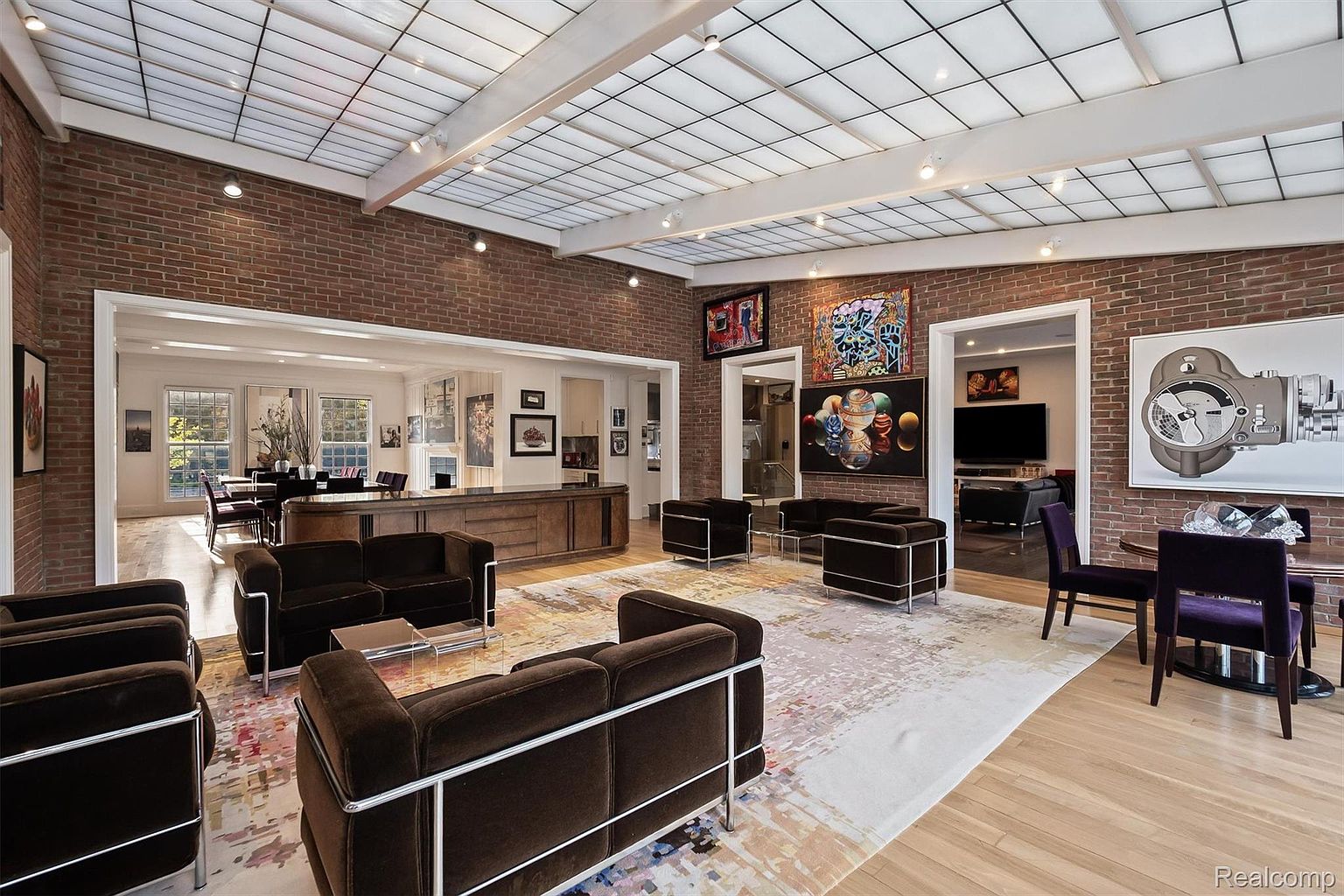 This is an interior shot of a spacious living room featuring a brick accent wall and a unique skylight ceiling. The room is furnished with modern, dark brown sofas with chrome frames, arranged around a light-colored rug with abstract patterns. Artwork adorns the walls, and there are open doorways leading to other rooms, suggesting an open-concept design.