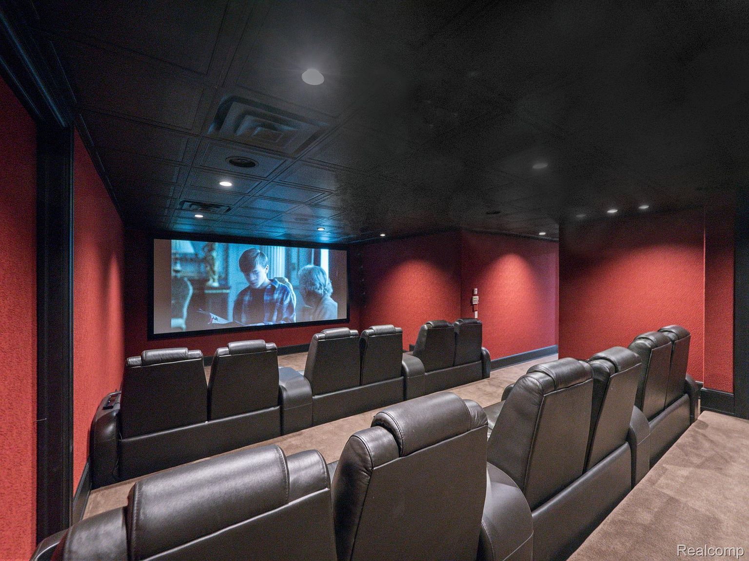 This is a home theater room featuring multiple rows of leather recliners facing a large screen displaying a movie. The walls are a textured red, and the ceiling is a dark, patterned material with recessed lighting. The room is designed for an immersive cinematic experience.