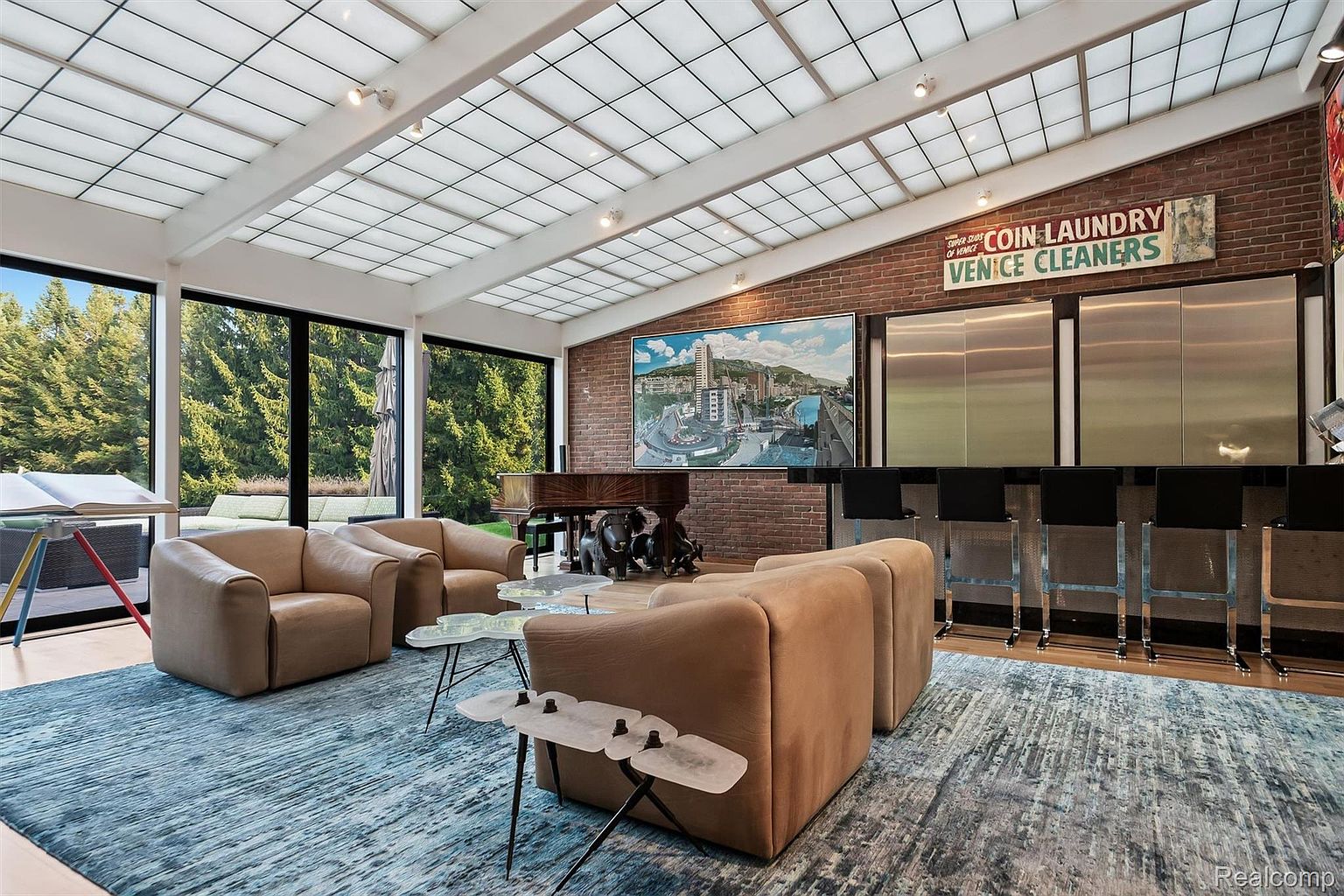 This is a spacious and modern living room featuring a unique skylight ceiling, brick accent wall, and a stylish bar area. The room is furnished with comfortable-looking tan armchairs and a distinctive blue and gray rug, creating a welcoming and sophisticated atmosphere. A grand piano sits against the brick wall, adding a touch of elegance.