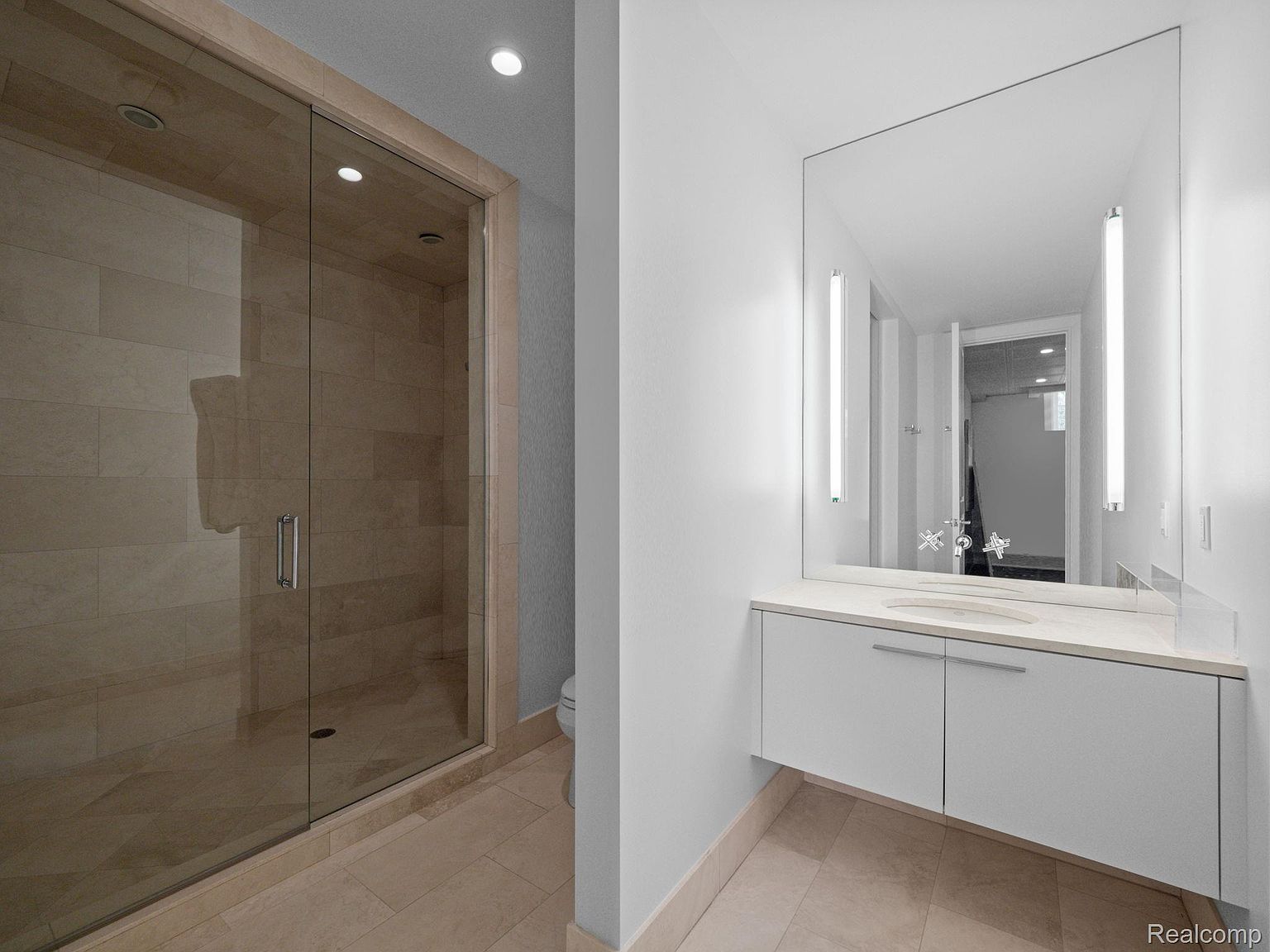 This is a modern primary bathroom featuring a glass-enclosed shower with beige tiling and a sleek vanity with a large mirror and integrated lighting. The color palette is neutral, creating a clean and serene atmosphere. The perspective is from the doorway, showcasing the shower and vanity area.