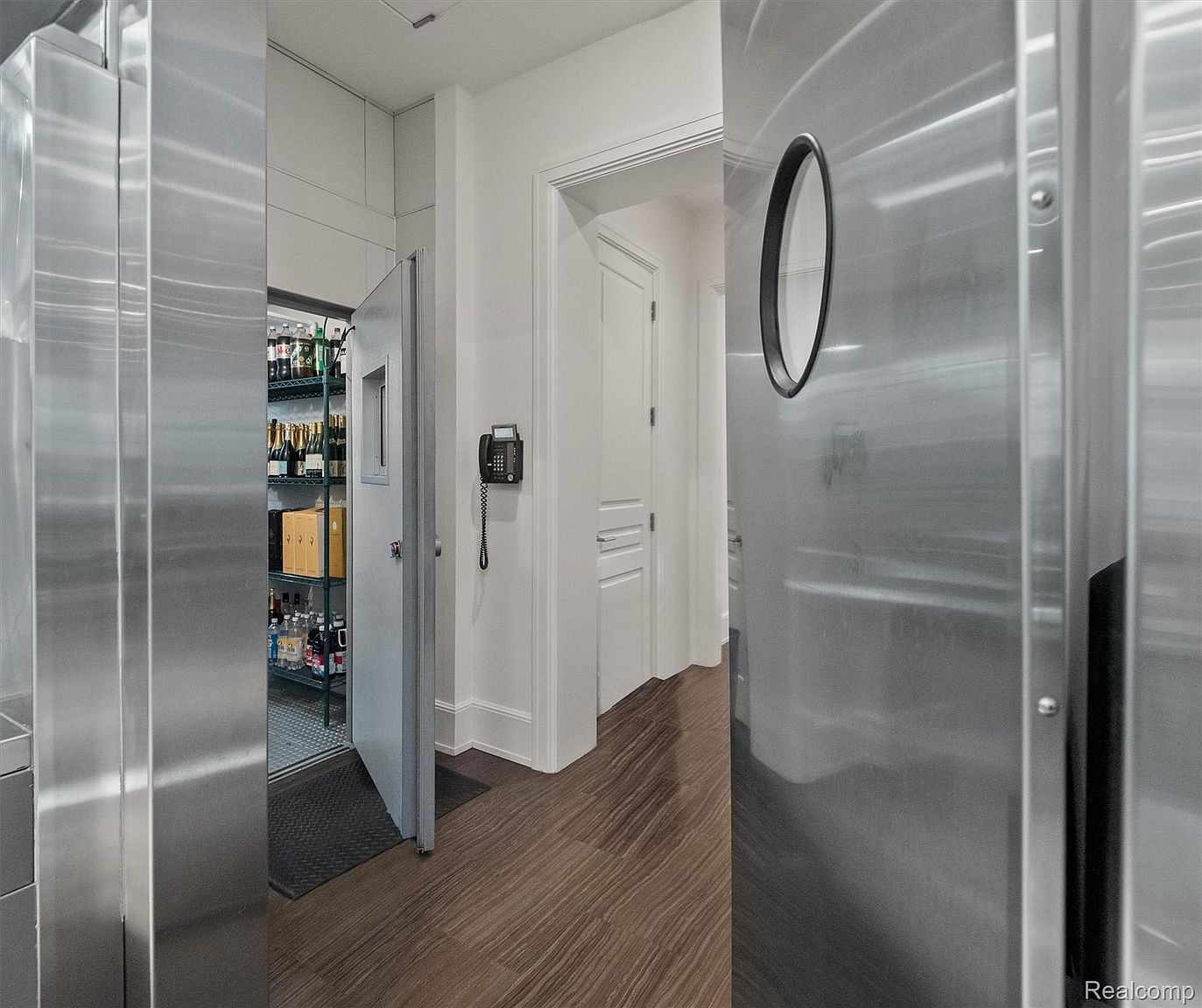 This image showcases a wine cellar with stainless steel shelving stocked with various wine bottles and boxes. The cellar door is partially open, revealing a glimpse of the interior, while a stainless steel door with a circular window is visible on the right. The flooring is a dark wood-like material, and a black wall-mounted telephone is seen in the hallway outside the cellar.