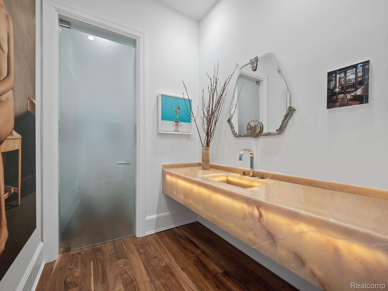 This is a modern bathroom featuring a unique, illuminated marble countertop with an integrated sink. An octagonal mirror hangs above the sink, complemented by decorative branches in a vase. The room is painted in a clean white, with wood flooring adding warmth and texture. A frosted glass door is on the left.