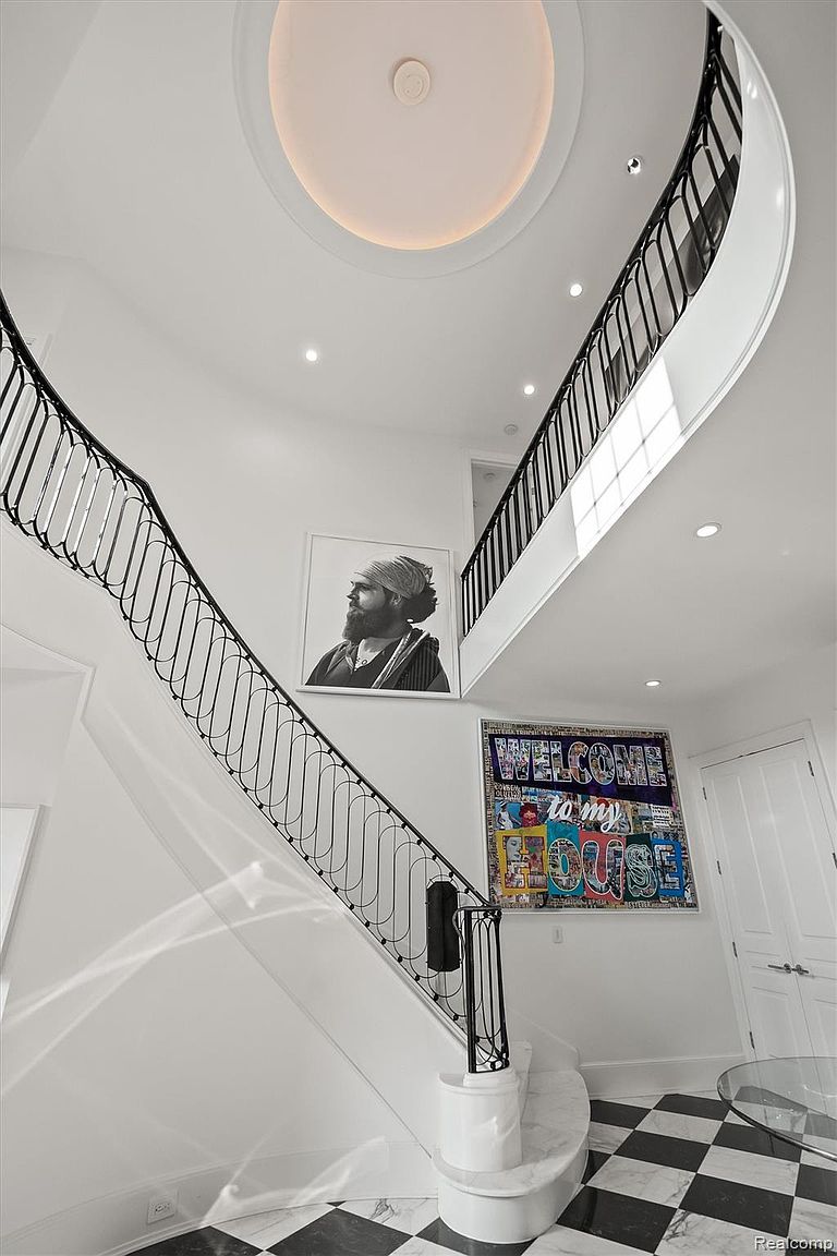 This is an interior shot of a grand hallway featuring a staircase with black wrought iron railings and white risers. The walls are painted white, and artwork adorns the walls, including a large portrait and a colorful 'Welcome to my House' piece. The flooring is a striking black and white checkered pattern, and a circular recessed light fixture is in the ceiling.
