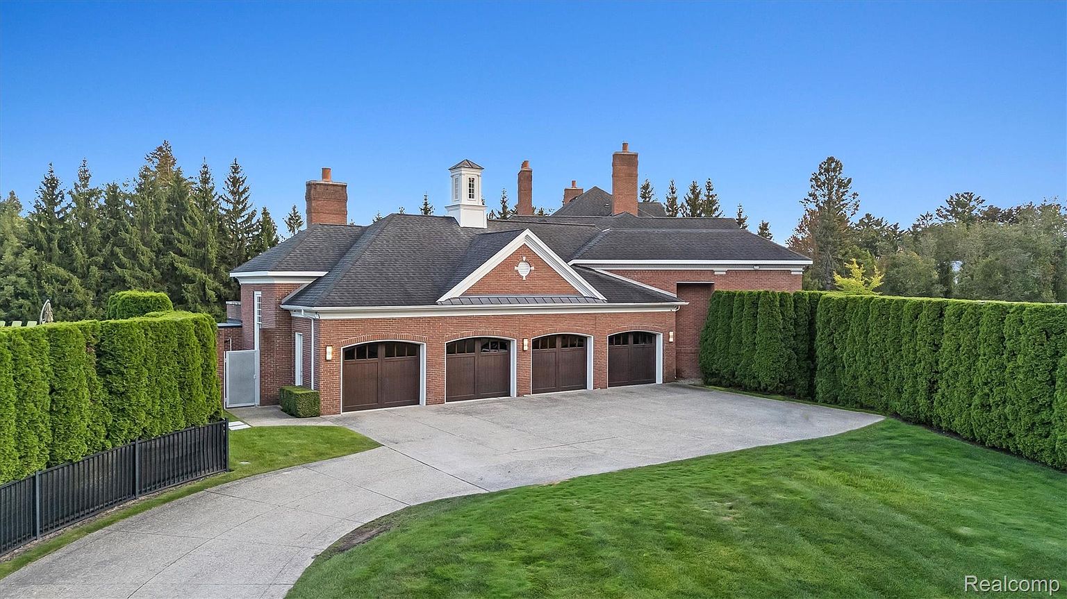 This is a front exterior view of a stately brick home featuring a prominent three-car garage with dark wood doors. The house is surrounded by manicured lawns and tall, dense hedges, providing privacy. The roof is dark and accented by multiple chimneys and a central cupola, adding to the home's architectural interest.