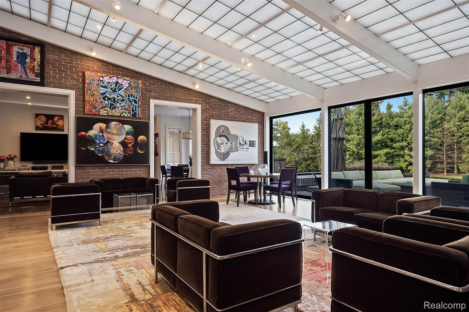 This is a spacious living room featuring a brick accent wall adorned with modern art, complemented by a skylight ceiling that floods the room with natural light. The room is furnished with contemporary brown sofas with metal frames, arranged around a patterned area rug, creating a stylish and inviting atmosphere. Large windows offer views of the lush greenery outside, enhancing the connection to nature.