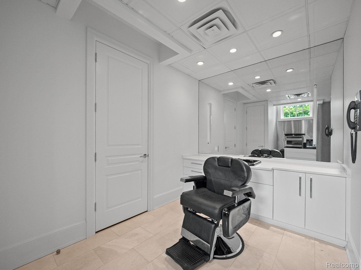 This interior shot showcases a well-lit barber room featuring a black barber chair as the focal point. The room is designed with a clean, modern aesthetic, characterized by white walls, cabinetry, and a large mirror that enhances the sense of space. The flooring appears to be a light-colored tile, contributing to the room's bright and airy feel.
