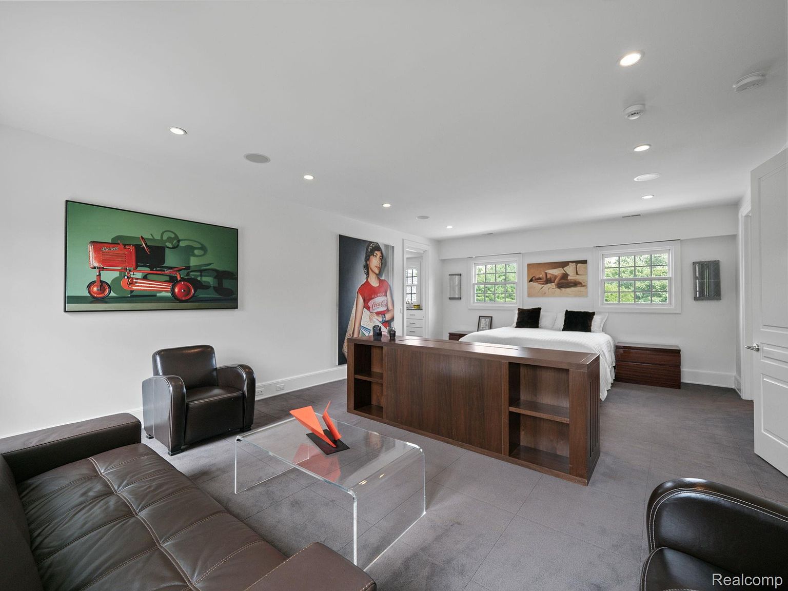 This is a spacious primary bedroom featuring a modern design with a gray carpet and white walls. The room includes a king-size bed with white linens and black pillows, flanked by wooden nightstands. A unique wooden room divider with built-in shelving separates the sleeping area from a seating area with a brown leather sofa and armchairs, centered around a clear acrylic coffee table. Artwork adorns the walls, and recessed lighting illuminates the space, creating a luxurious and comfortable atmosphere.