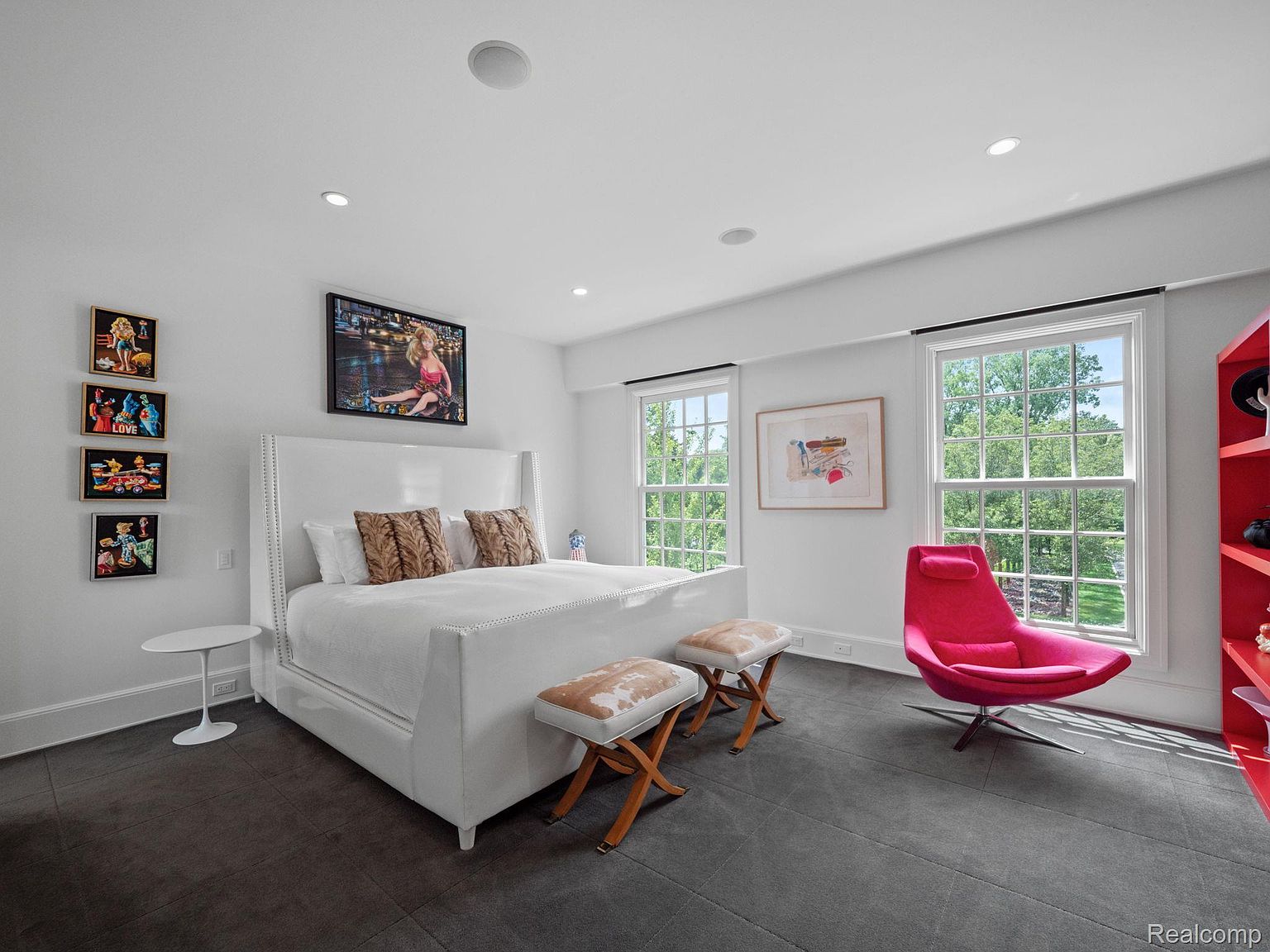 This is a bright and modern primary bedroom featuring a large white bed with decorative pillows, flanked by a white side table and two upholstered stools. The room is well-lit with natural light from two large windows, complemented by recessed lighting. Artwork adorns the walls, and a vibrant pink chair adds a pop of color against the dark flooring, creating a stylish and inviting space.