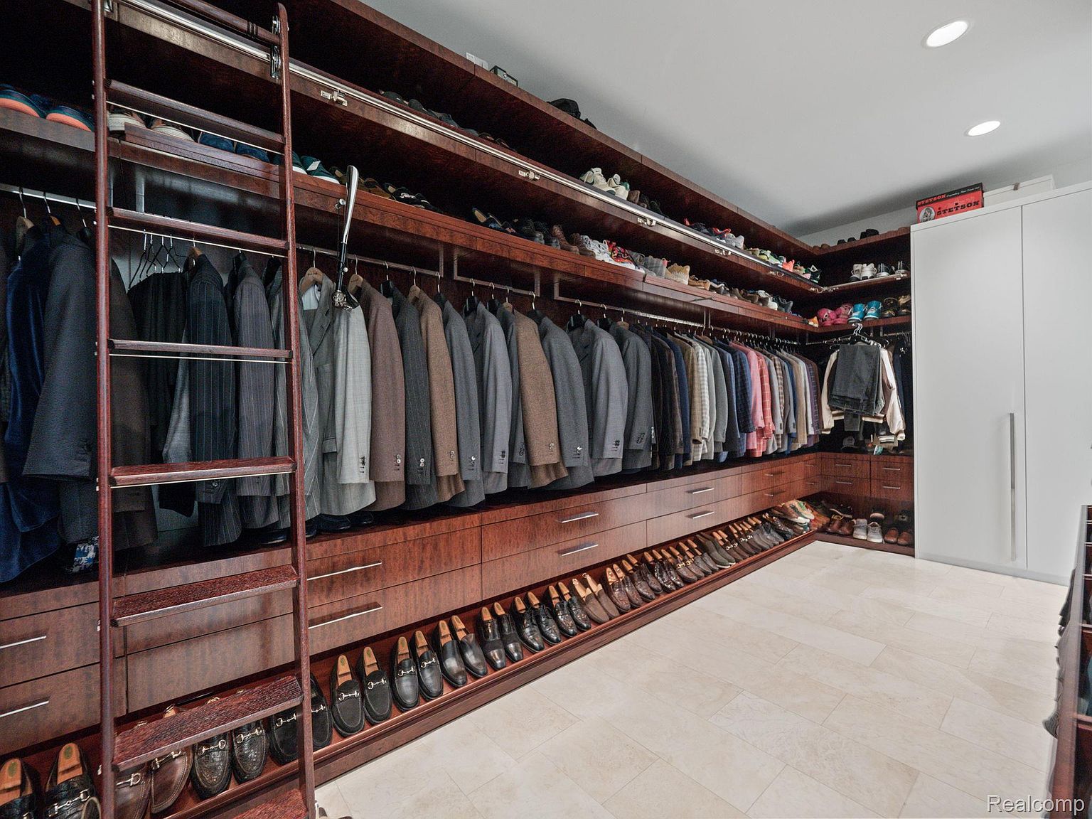 This is a well-organized walk-in closet featuring dark wood cabinetry and ample storage space. The closet includes hanging rods for suits and shirts, drawers for folded items, and shoe racks along the bottom. A rolling ladder provides access to higher shelves, and the overall design is luxurious and functional.
