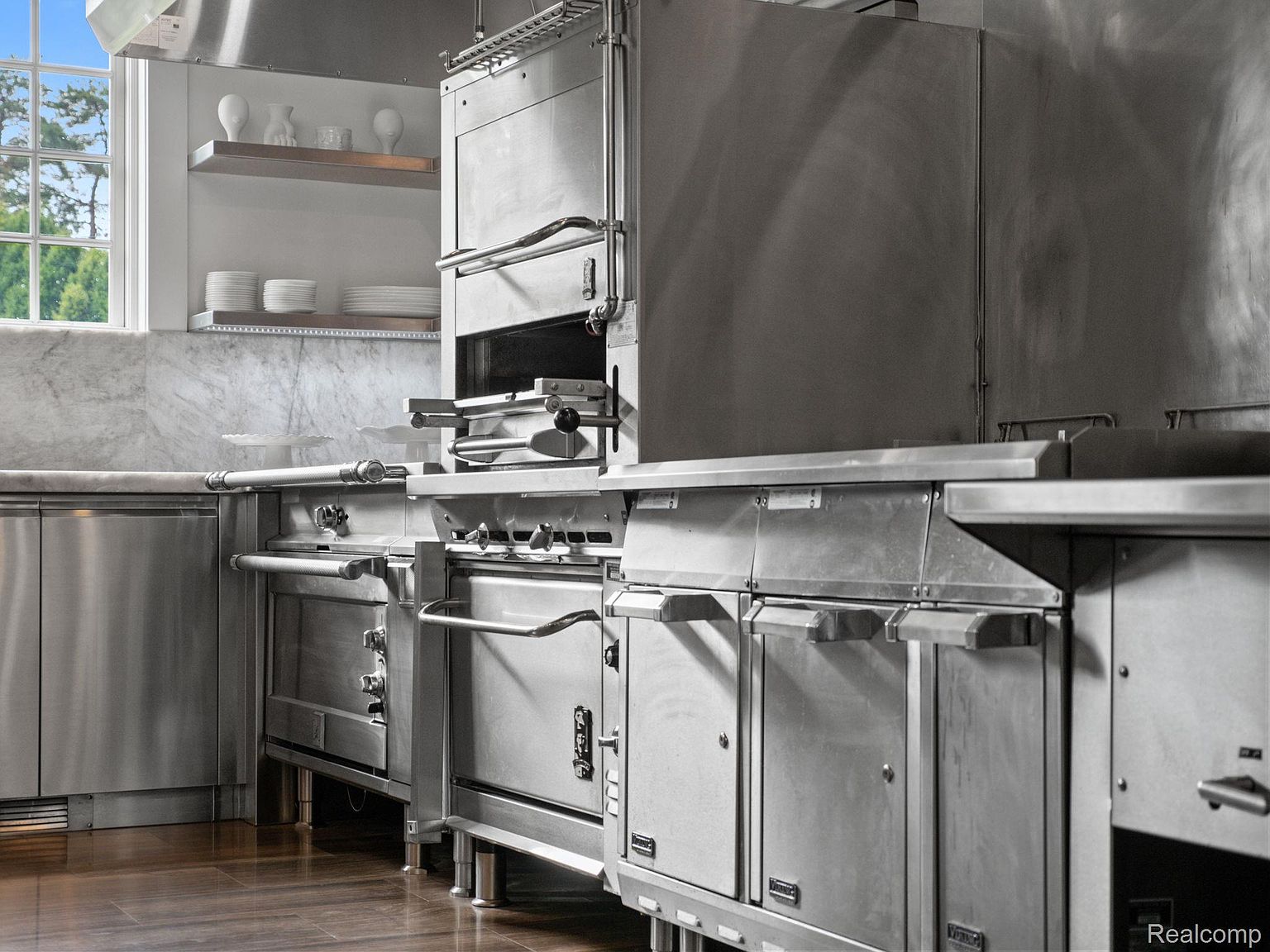 This is a professional-grade kitchen featuring stainless steel appliances and cabinetry. The kitchen is well-lit, with a window providing natural light. The overall impression is one of functionality and high-end quality, suitable for a serious cook.