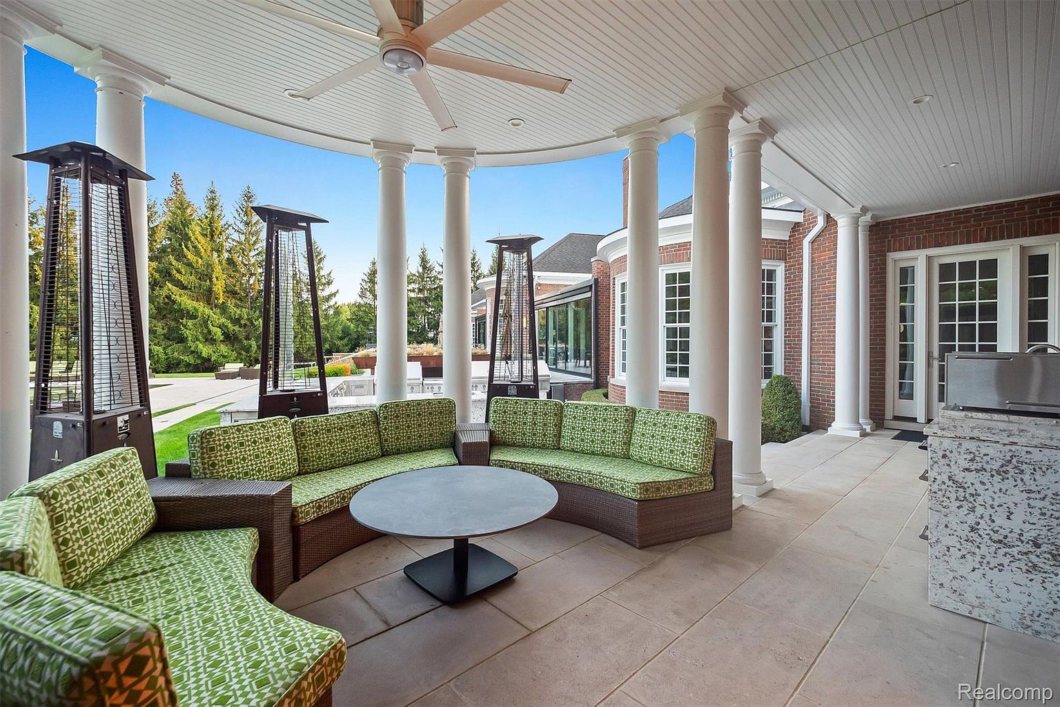 This image showcases a luxurious outdoor patio area, featuring a curved seating arrangement with green patterned cushions around a round table. The patio is covered by a structure supported by white columns and a ceiling fan, providing shade and comfort. Tall, modern patio heaters add warmth and ambiance, while the backdrop includes a well-manicured lawn and a glimpse of the brick house, creating an inviting and upscale outdoor living space.