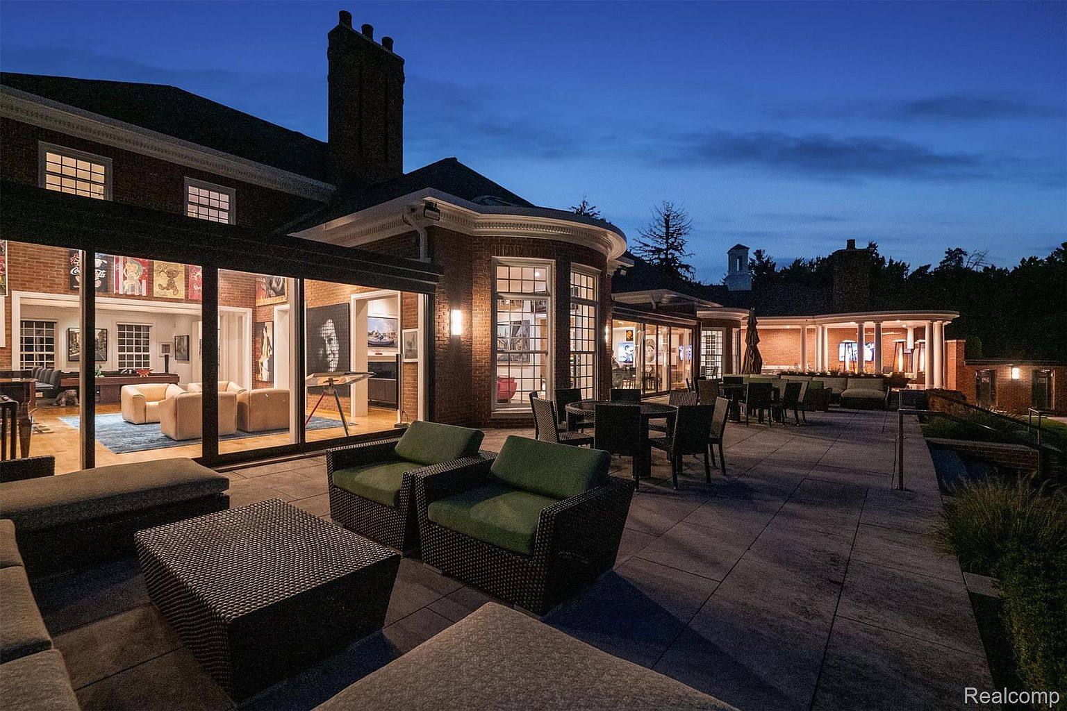 This image showcases a luxurious patio area at dusk, featuring comfortable outdoor seating arrangements and dining tables. The patio is connected to a grand brick building with large windows offering glimpses into the interior, which appears to be a living area with stylish furniture and artwork. The overall impression is one of sophisticated outdoor living and elegant design.