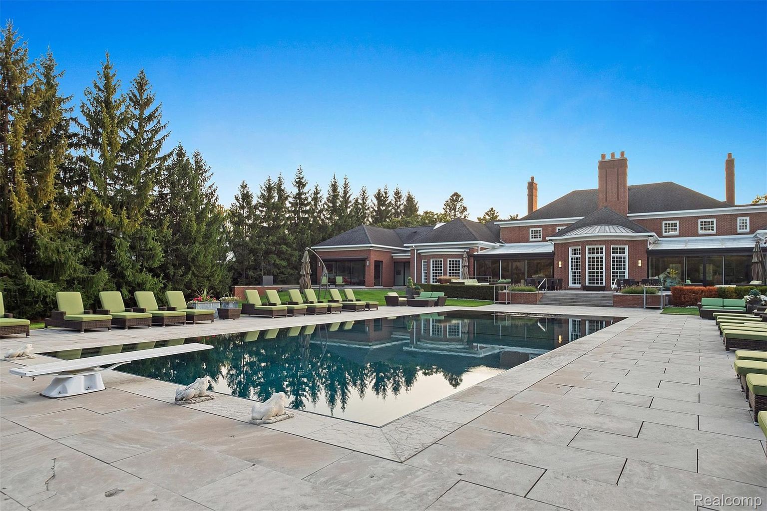 This image showcases a luxurious backyard featuring a large rectangular pool with a diving board and elegant stone coping. The pool reflects the surrounding trees and the impressive brick mansion in the background, complete with multiple chimneys and a sunroom. Lounge chairs are arranged around the pool, creating an inviting and relaxing atmosphere.
