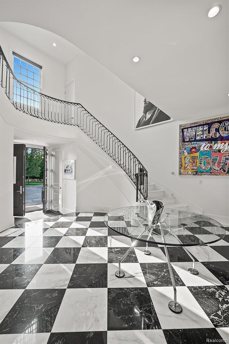This is an interior shot of a grand foyer featuring a black and white checkered marble floor, a curved staircase with a black wrought iron railing, and white walls adorned with modern art. A round glass table with a unique chrome base sits in the foreground, adding a contemporary touch to the classic design. The open doorway leads to a glimpse of the exterior, enhancing the sense of spaciousness and light.