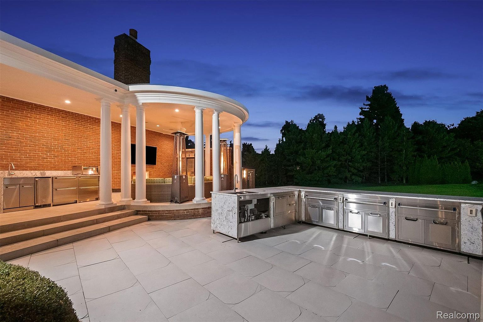 This is a rear exterior view of a luxurious patio area featuring an outdoor kitchen with stainless steel appliances and marble countertops. The patio is paved with large, light-colored tiles and is partially covered by a colonnaded structure with a brick wall backdrop. The scene is set at dusk, with a clear blue sky and mature trees in the background, creating an inviting and upscale outdoor living space.