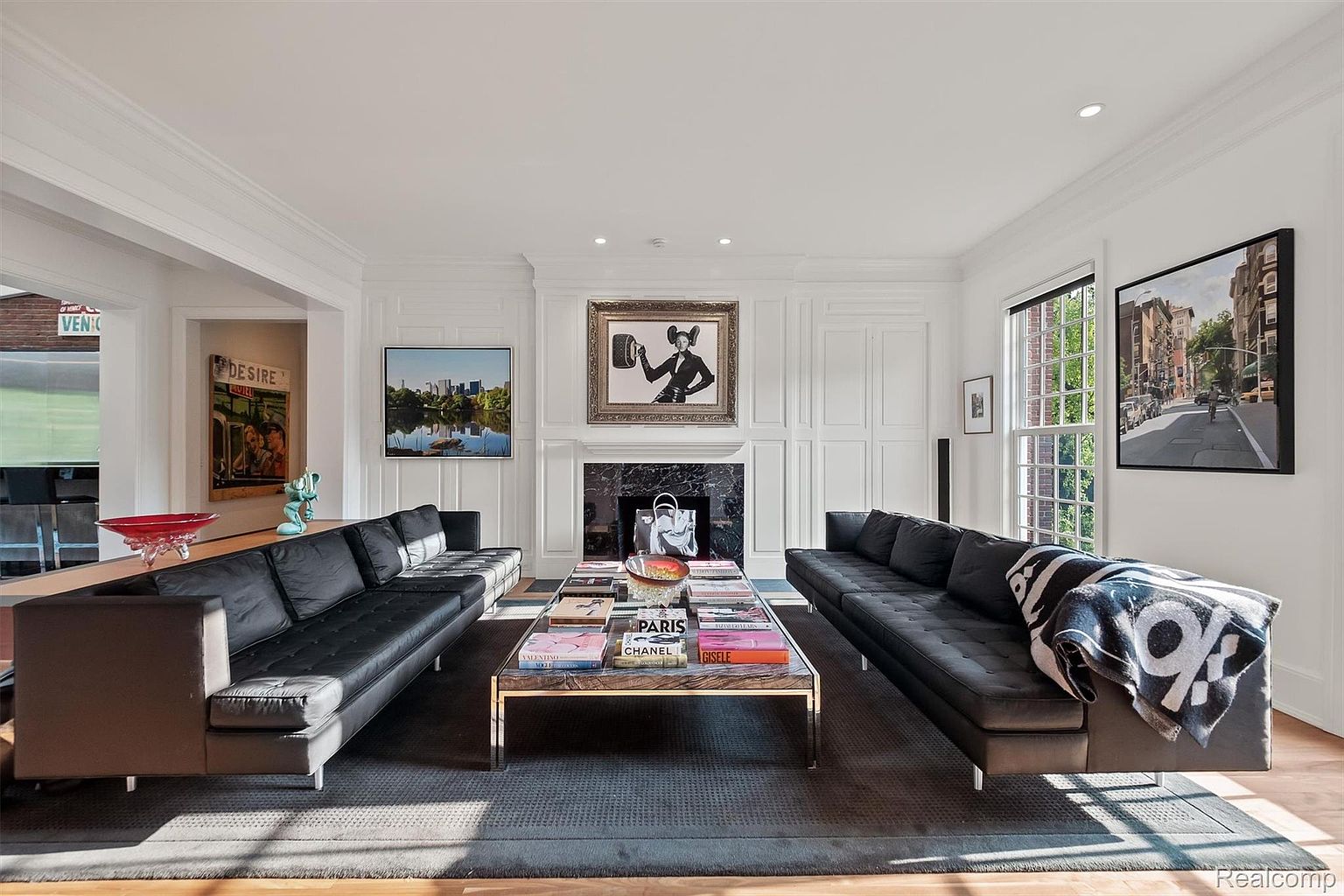 This is an interior shot of a living room featuring two black leather sofas facing each other, separated by a modern coffee table with books. The room has white walls with wainscoting, artwork above the fireplace and on the adjacent walls, and a large window providing natural light. The overall impression is sophisticated and contemporary.