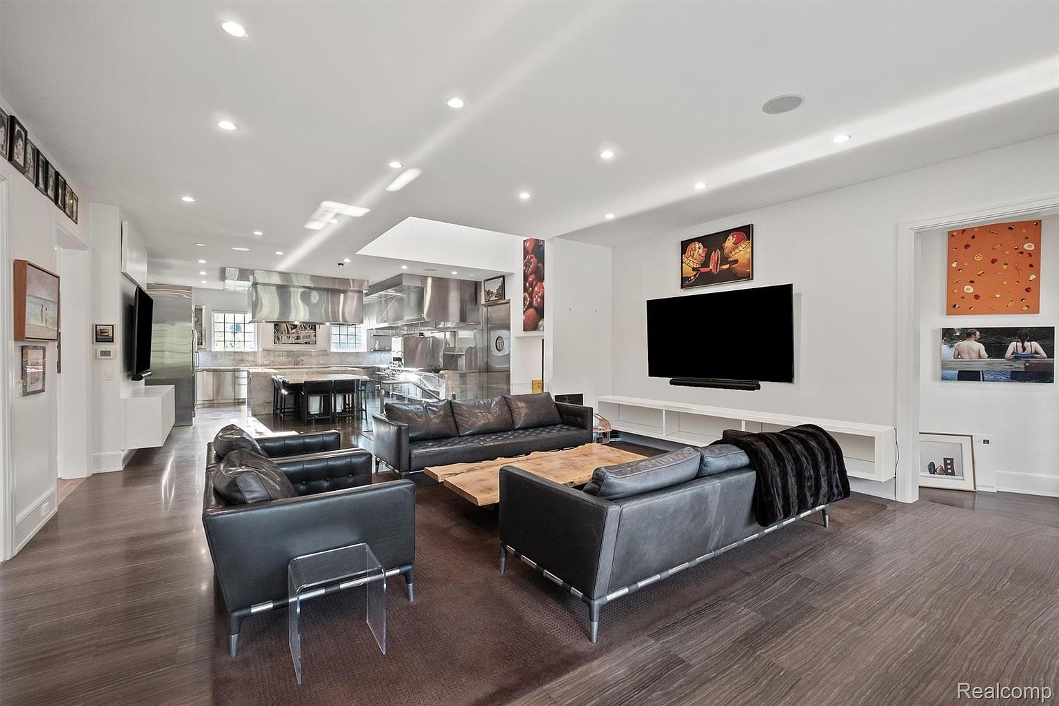 This is an interior shot of a modern living room that seamlessly blends into a high-end kitchen. The living room features dark hardwood floors, black leather sofas, and a large flat-screen TV mounted on the wall above a white console. The kitchen boasts stainless steel appliances and a large island with bar stools, creating an open and inviting space.