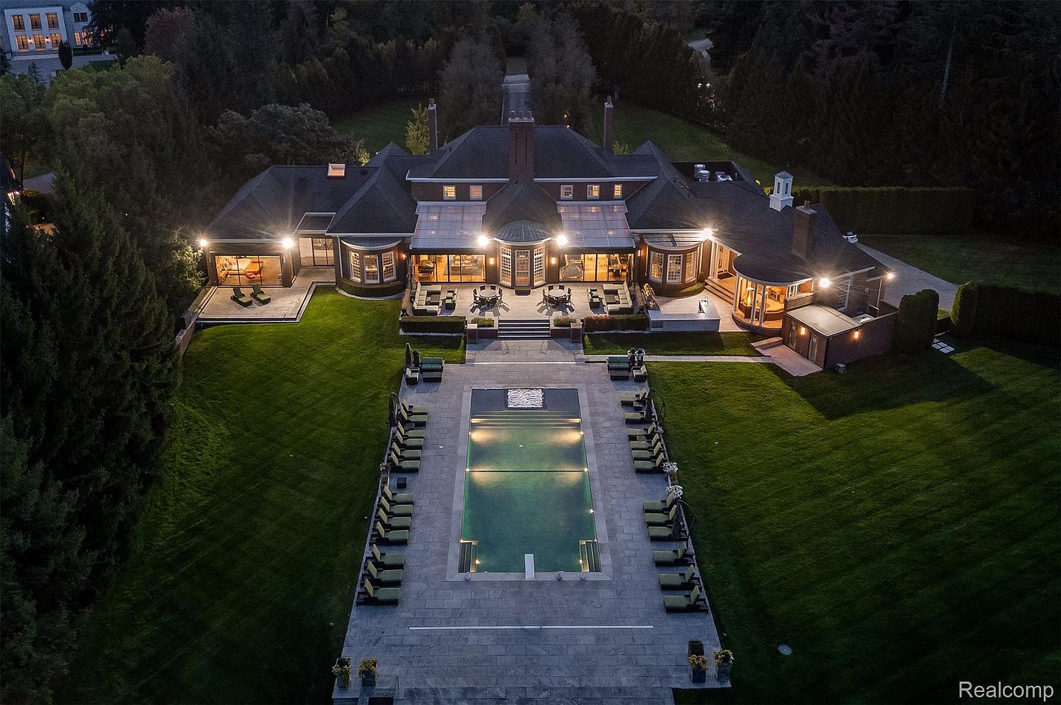 This aerial view showcases a luxurious estate at night, featuring a sprawling mansion with a dark roof, illuminated windows, and manicured lawns. A large rectangular pool, surrounded by lounge chairs and a stone patio, serves as a focal point, while the overall impression is one of grandeur and sophisticated outdoor living.