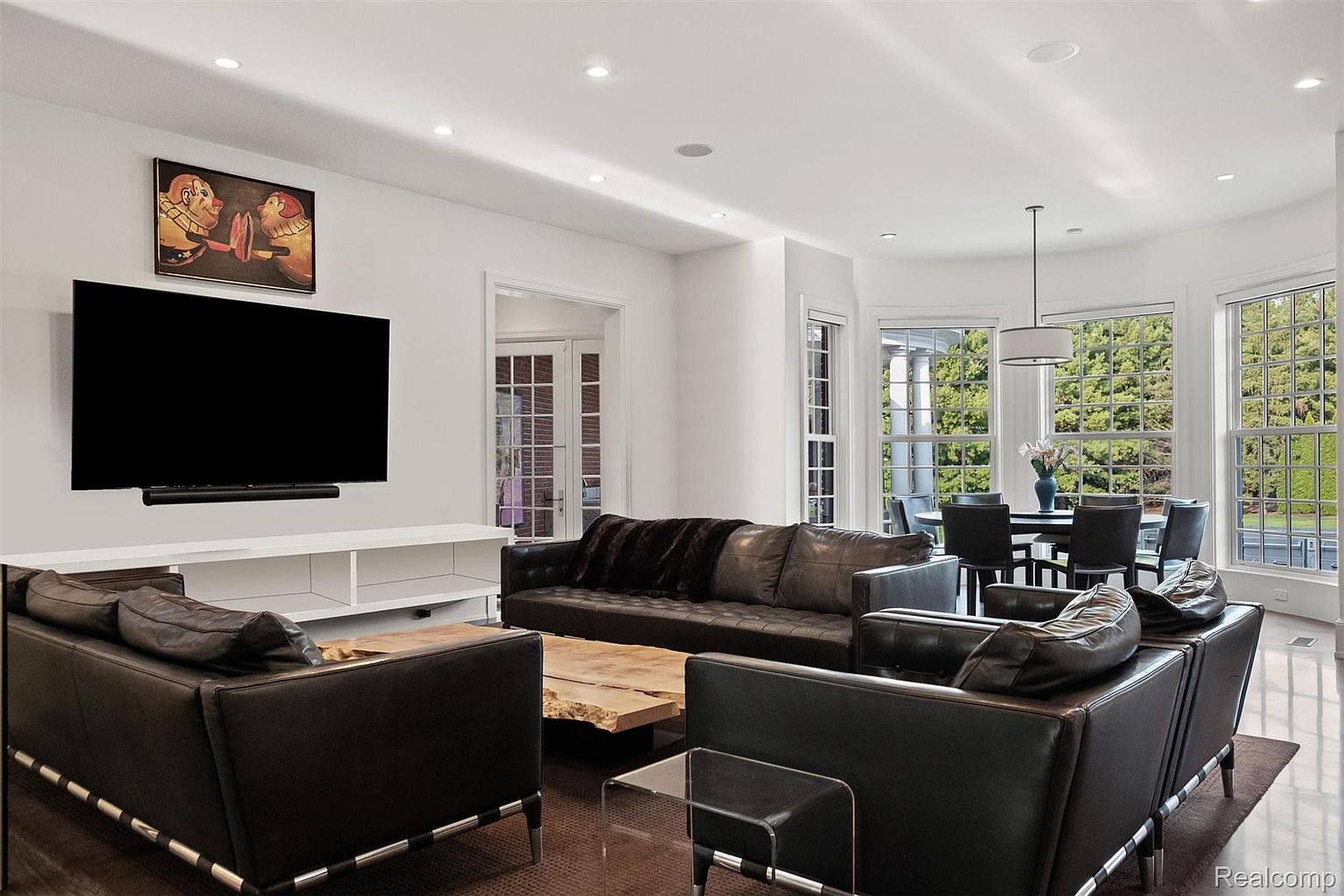 This is an interior shot of a modern living room and dining area. The living room features black leather sofas, a unique wooden coffee table, and a large television mounted above a white console. The dining area is visible through large windows, showcasing a round table and chairs, creating an open and inviting space.