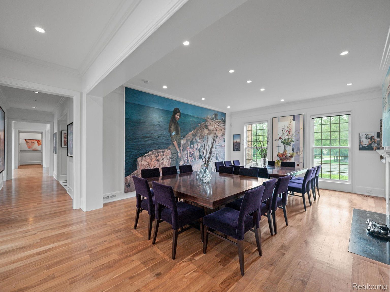 This is an interior shot of a formal dining room. The room features a large, dark wood square dining table surrounded by purple upholstered chairs. A large painting dominates one wall, while windows provide natural light. The hardwood floors and recessed lighting contribute to the room's elegant atmosphere.