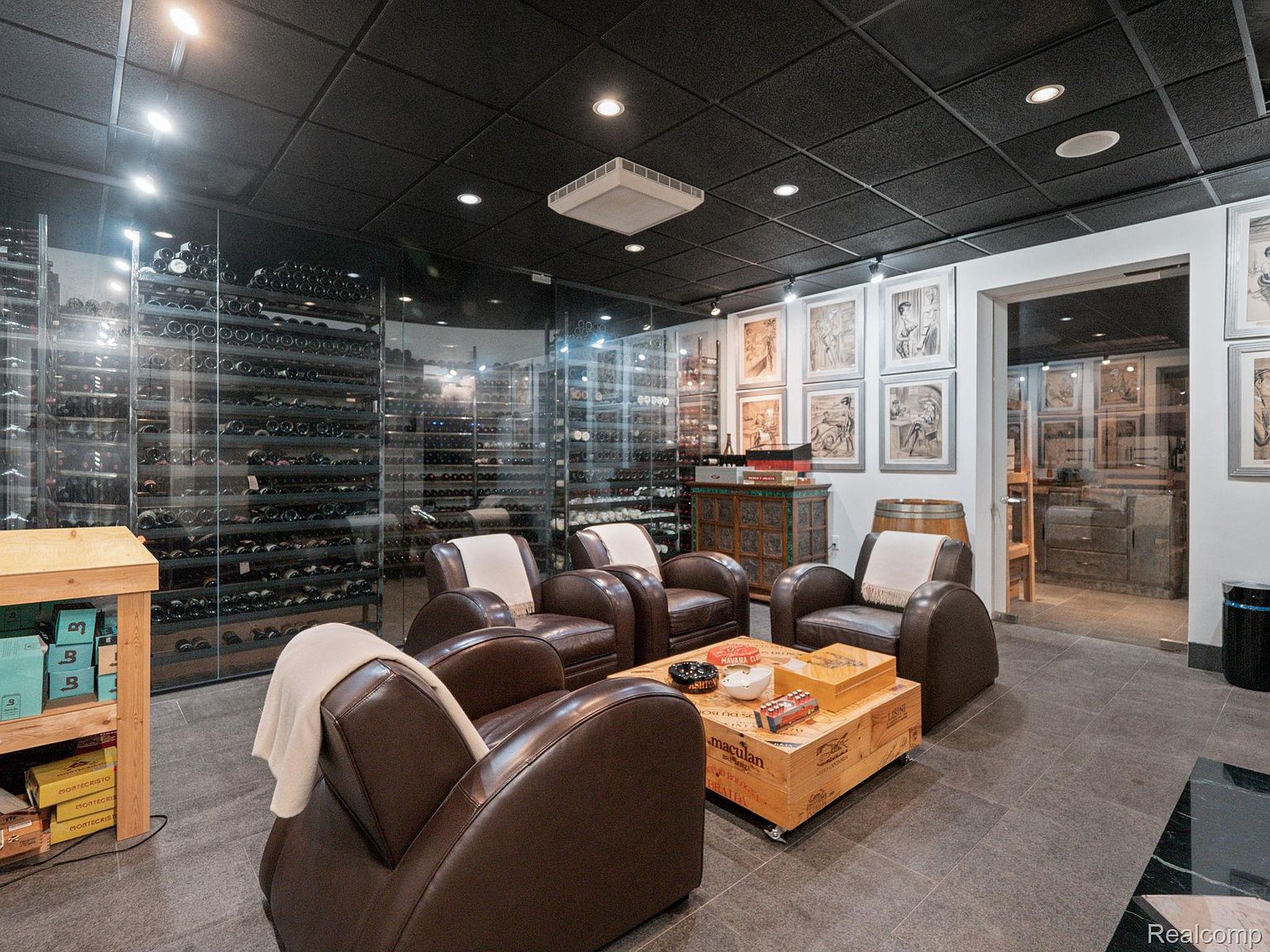 This is an interior shot of a sophisticated wine cellar. The room features extensive wine storage behind glass walls, comfortable leather armchairs arranged around a wooden crate coffee table, and artwork adorning the walls. The dark ceiling and tiled floor contribute to a luxurious and inviting atmosphere, perfect for wine tasting and relaxation.