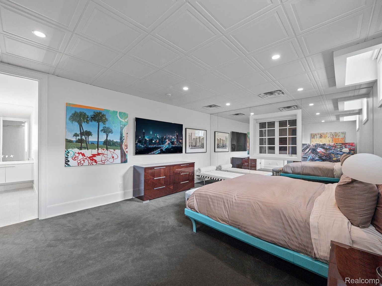 This is a primary bedroom featuring a large bed with a light blue frame and neutral bedding. The room has a modern aesthetic with a mirrored wall, recessed lighting, and artwork displayed above a wooden dresser. The flooring is a dark gray carpet, and there is a glimpse into an adjacent bathroom through an open doorway.