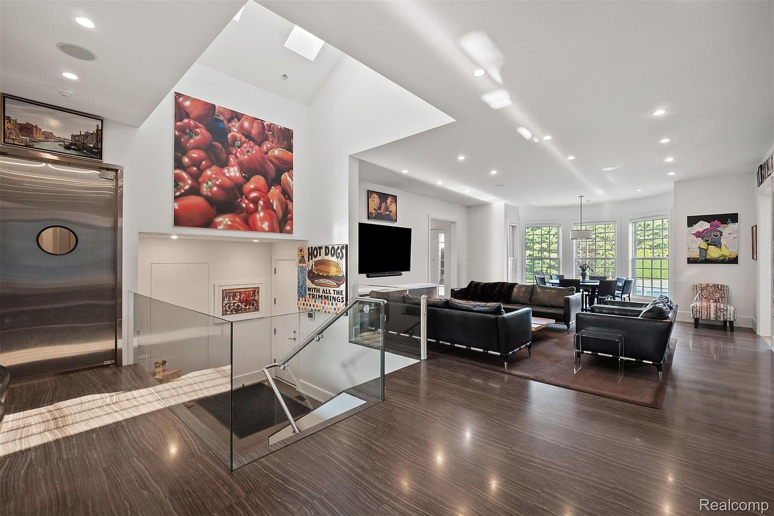 This is a spacious and modern living room featuring hardwood floors, recessed lighting, and a high ceiling with a skylight. The room is furnished with multiple dark leather sofas and a dining table set in the background. Artwork and a large television adorn the walls, adding character to the space. The perspective is from the entrance of the living room.
