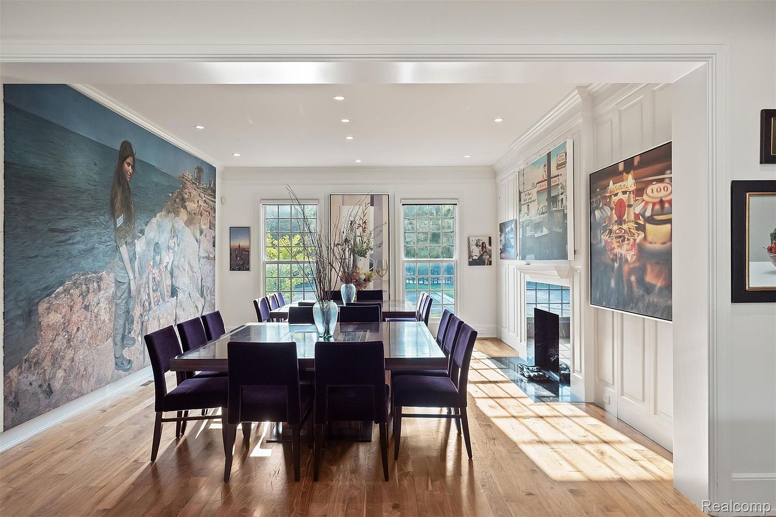 This is a well-lit dining room featuring a long, dark wood table with purple upholstered chairs. Large artwork adorns the walls, and natural light streams in through multiple windows. The room has a sophisticated and elegant feel, perfect for formal dining.