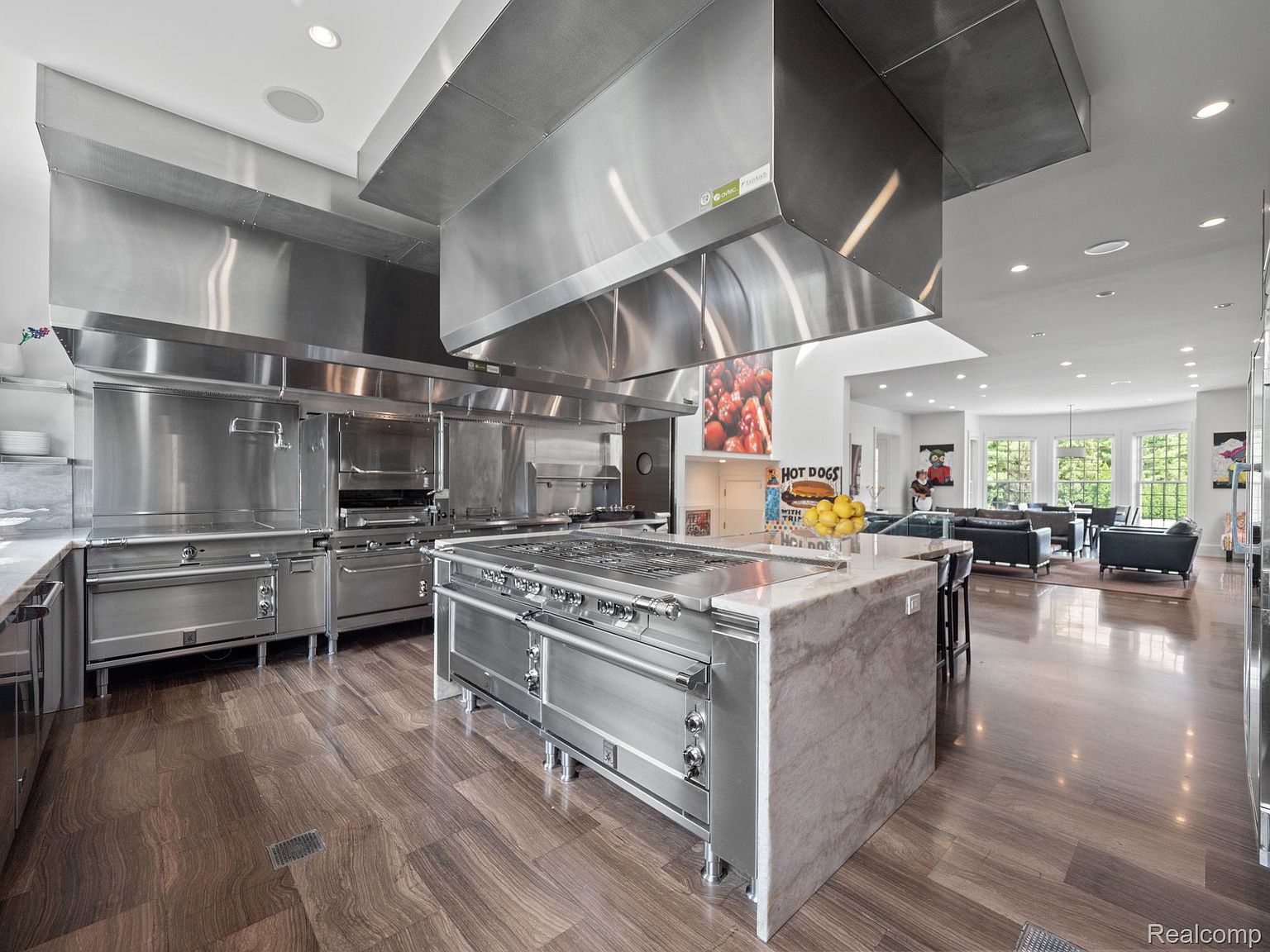 This is a professional-grade kitchen featuring stainless steel appliances, including a large range with a custom hood. The kitchen island has a marble countertop and the flooring is a dark wood. The space opens to a living area, creating an open-concept design.