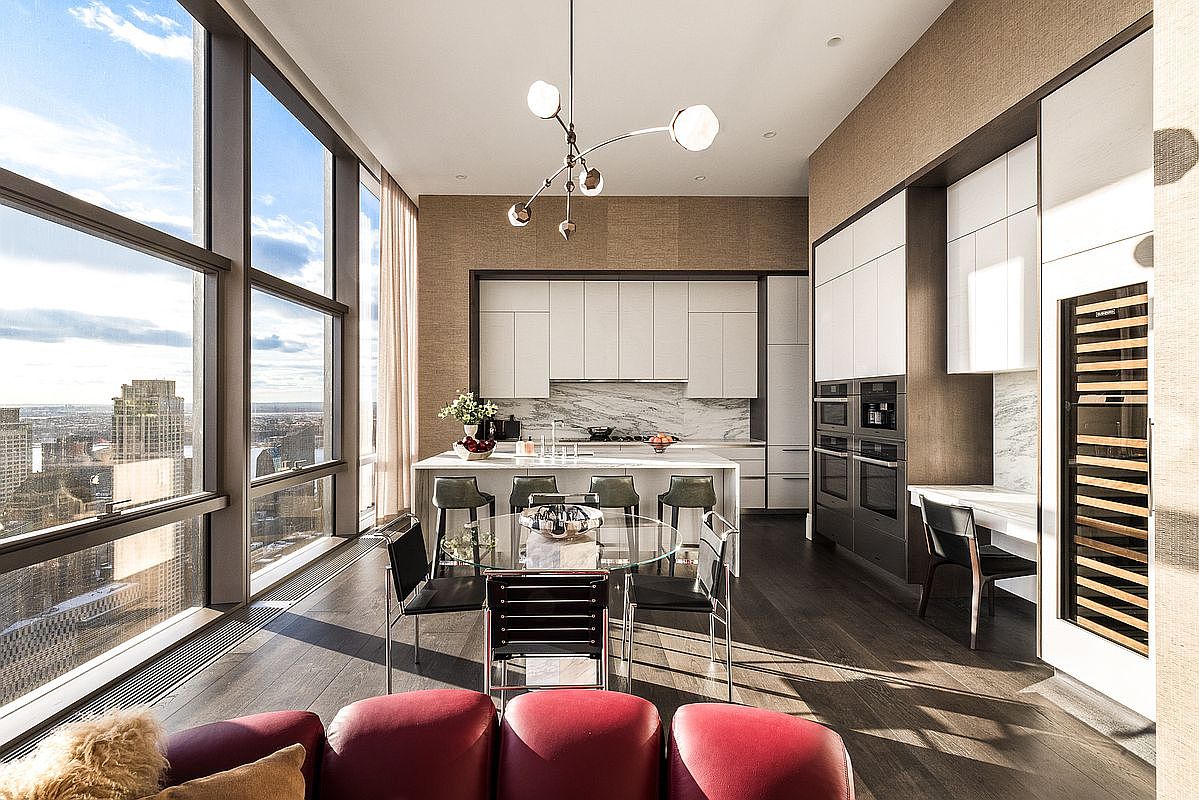 This sophisticated, high-rise kitchen features a sleek, modern design with white cabinetry, marble countertops, and a central island with bar seating. A glass dining table sits in the foreground, while floor-to-ceiling windows provide a breathtaking, expansive view of the city skyline. The space is illuminated by a contemporary chandelier, creating an elegant and airy atmosphere perfect for luxury urban living.