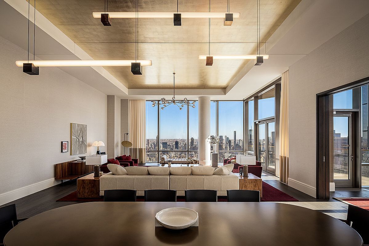 This expansive, high-end living room features a sophisticated open-concept layout with floor-to-ceiling windows that offer a breathtaking panoramic view of the city skyline. The space is anchored by a large, neutral-toned sectional sofa, complemented by elegant gold-leaf ceiling accents and modern linear pendant lighting. The perspective from the foreground dining area captures the luxurious scale and refined contemporary aesthetic of this urban penthouse.