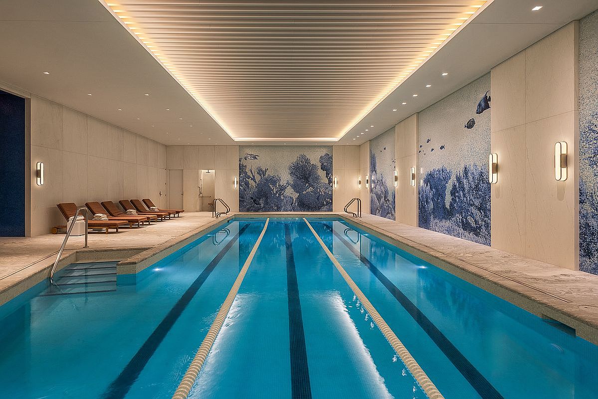 This luxurious indoor lap pool features a sleek, modern design with a striking blue mosaic mural depicting underwater coral and marine life. The space is illuminated by recessed lighting and a sophisticated slatted ceiling, while a row of wooden lounge chairs provides a relaxing area for residents. The perspective is a wide, symmetrical shot from the pool deck, emphasizing the clean lines and high-end finishes of this premium amenity.