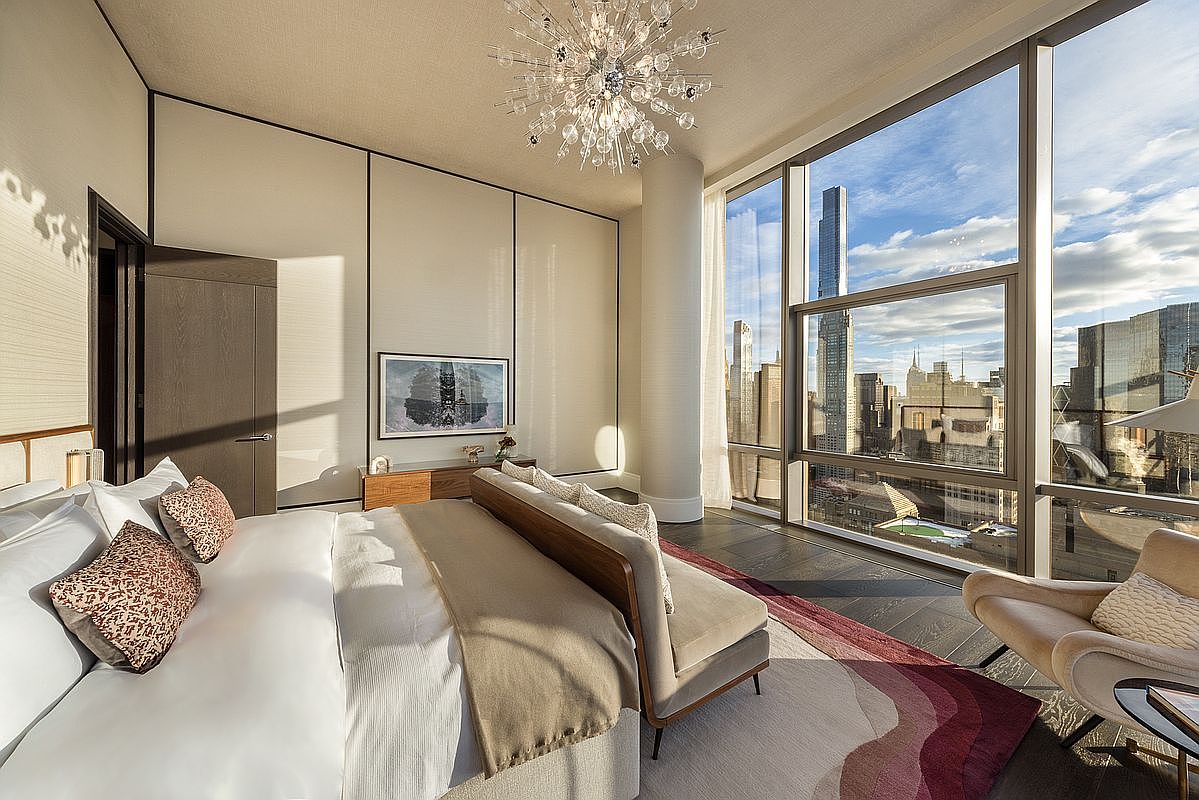 This luxurious primary bedroom features floor-to-ceiling windows that offer a breathtaking, panoramic view of the New York City skyline. The room is elegantly appointed with a plush bed, a modern chandelier, and a sophisticated color palette of neutrals and deep reds. The combination of high-end finishes and the dramatic urban backdrop creates an atmosphere of refined, contemporary opulence.