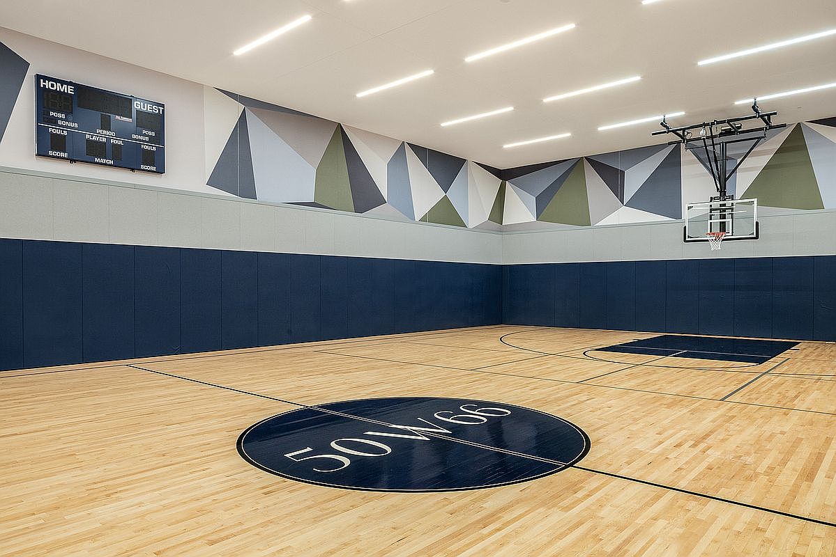 This indoor basketball court features a polished hardwood floor with a prominent circular logo in the center and a dark blue painted key area. The walls are adorned with a modern geometric mural in shades of blue, grey, and olive, complemented by protective blue wall padding. A digital scoreboard and a professional-grade basketball hoop complete the space, creating a clean, high-end recreational facility.