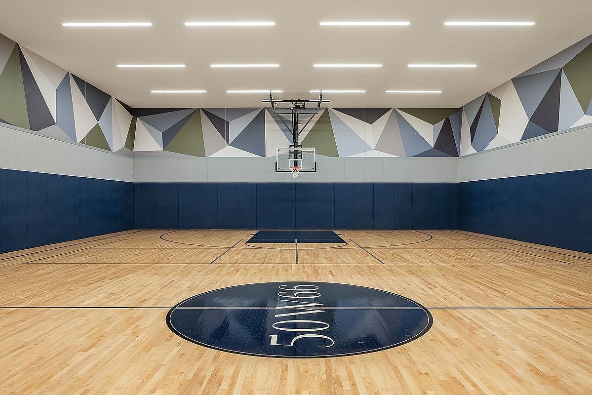 This indoor basketball court features a polished hardwood floor with a prominent '50W66' logo at center court. The walls are adorned with a modern, geometric-patterned mural in shades of blue, grey, and olive, complemented by a dark blue padded wainscoting. The space is brightly lit by recessed linear ceiling lights, creating a clean, professional, and inviting recreational environment.