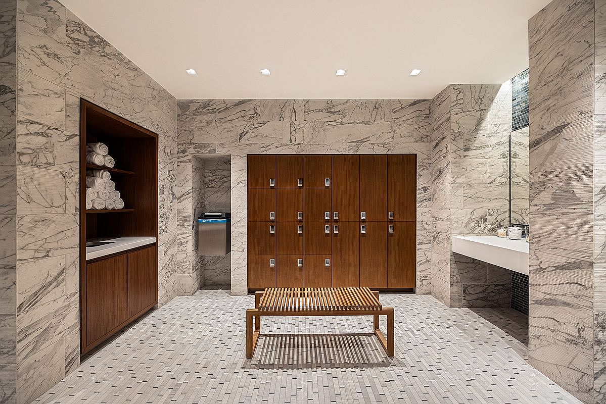 This sophisticated locker room facility features high-end finishes, including floor-to-ceiling marble wall cladding and a sleek, slatted wood bench as the central focal point. The space is equipped with warm-toned wooden lockers, a dedicated towel storage area, and a clean, minimalist vanity section. The overall aesthetic is one of luxury and tranquility, typical of a high-end residential or club amenity space.