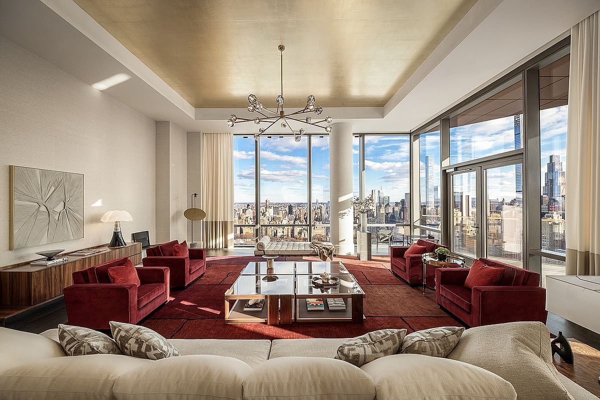This luxurious, high-rise living room features a sophisticated design with a striking gold-leaf ceiling and floor-to-ceiling windows that offer expansive, panoramic city views. The space is anchored by a deep red area rug and plush velvet armchairs, complemented by a large, modular cream-colored sofa in the foreground. A modern, multi-tiered glass coffee table and a unique sputnik-style chandelier serve as elegant focal points, creating an atmosphere of refined urban opulence.