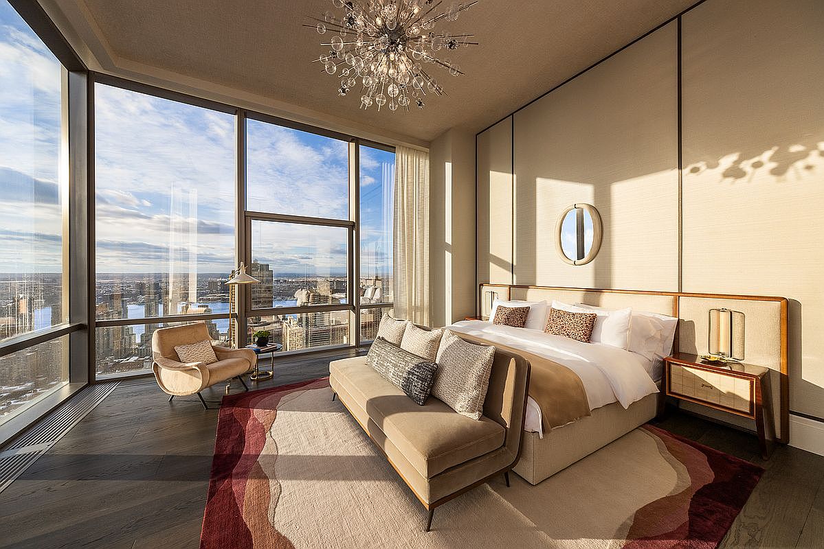 This luxurious primary bedroom features floor-to-ceiling windows that offer a breathtaking, panoramic view of the city skyline. The room is elegantly furnished with a plush bed, a sophisticated bench at the foot, and a stylish armchair, all anchored by a contemporary area rug. A stunning starburst chandelier serves as the centerpiece, casting a warm, inviting glow across the neutral-toned walls and high-end finishes.