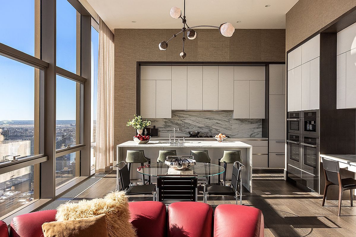 This sophisticated, high-rise kitchen features a sleek, modern design with white cabinetry, a marble-topped island, and integrated stainless steel appliances. A glass dining table sits in the foreground, while floor-to-ceiling windows offer a breathtaking, expansive view of the city skyline. The space is illuminated by a unique, sculptural chandelier, creating an atmosphere of luxury and refined urban living.