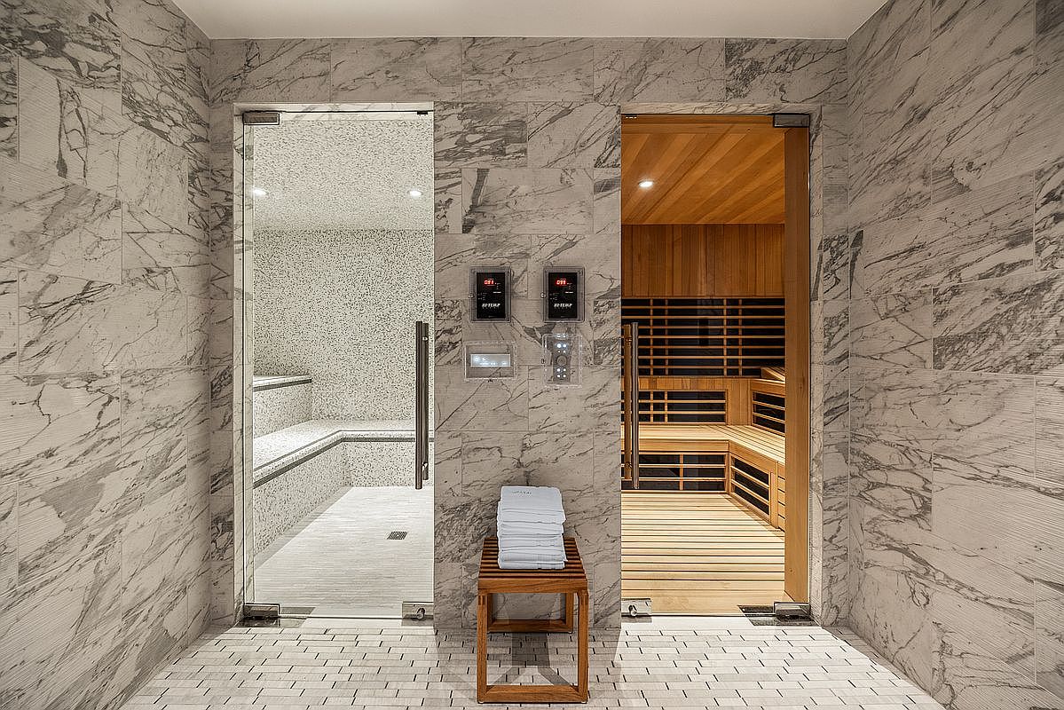 This luxurious spa area features a side-by-side steam room and dry sauna, both enclosed by sleek glass doors and framed by elegant, light-veined marble tile walls. A wooden bench holding a stack of fresh white towels sits centered between the two rooms, creating a balanced and inviting focal point. The space exudes a high-end, tranquil atmosphere typical of a premium residential or hotel wellness facility.
