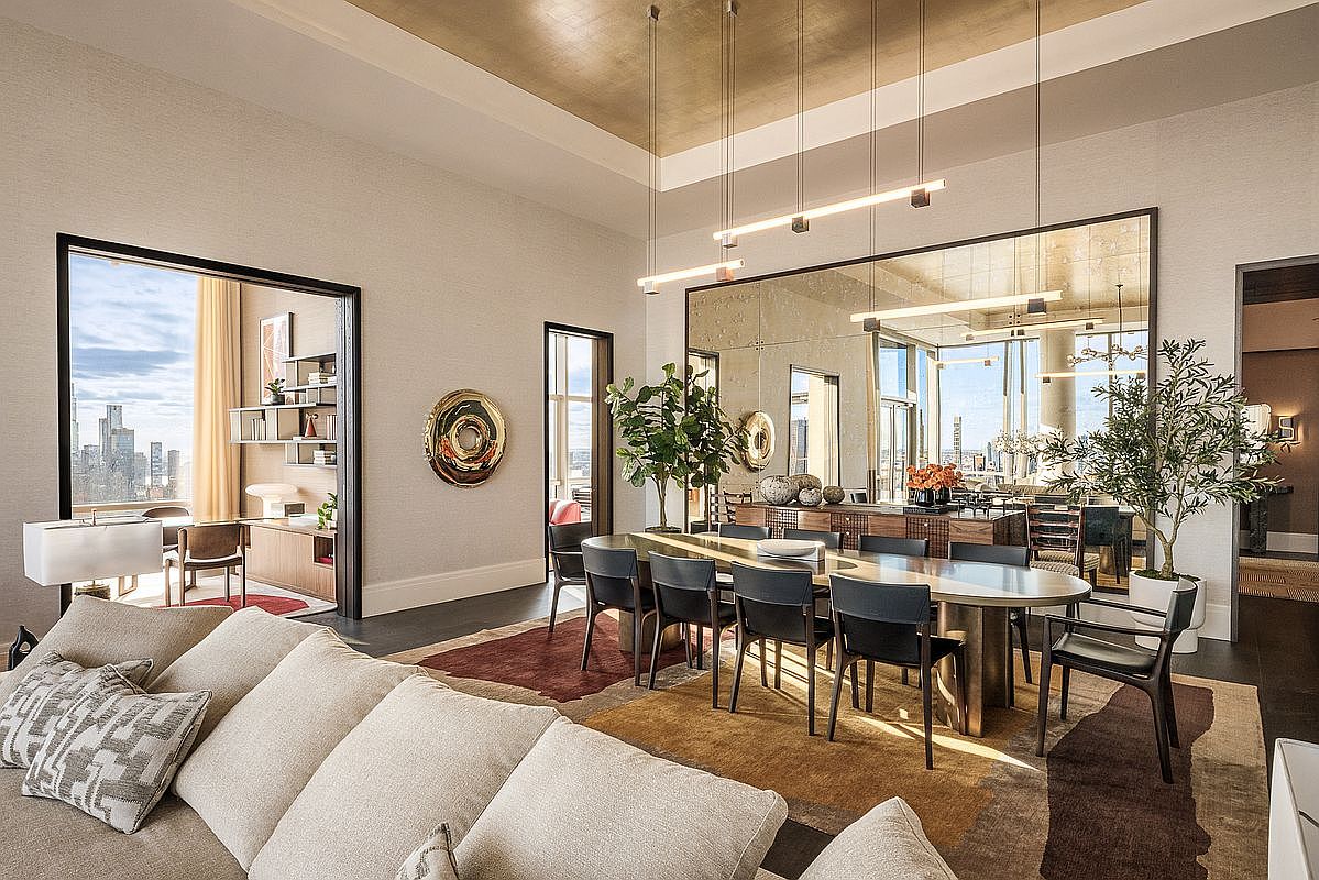 This sophisticated open-concept dining area features a sleek, oval-shaped table surrounded by modern black chairs, set against a large, reflective wall mirror that enhances the sense of space. The room is anchored by a textured, multi-toned area rug and illuminated by a minimalist linear pendant light fixture, while a nearby office space is visible through an open doorway. The overall aesthetic is one of refined luxury, characterized by high ceilings, warm metallic accents, and a seamless blend of contemporary design and urban views.