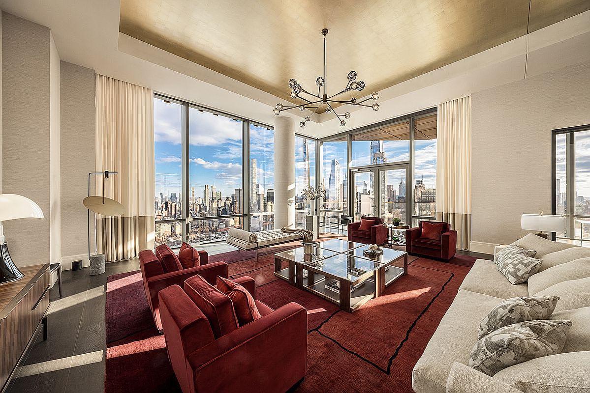 This luxurious, high-rise living room features floor-to-ceiling windows that offer a breathtaking panoramic view of the city skyline. The space is anchored by a deep red area rug, plush velvet armchairs, and a large, comfortable sofa, all illuminated by a striking modern chandelier suspended from a gold-leaf ceiling. The sophisticated design blends contemporary elegance with dramatic architectural elements, creating an atmosphere of refined urban opulence.