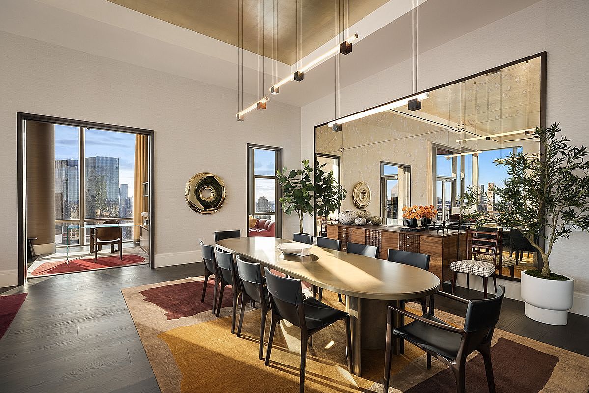 This sophisticated dining room features a large, oval-shaped table surrounded by sleek black leather chairs, set against a backdrop of a grand, floor-to-ceiling mirror that enhances the sense of space. The room is illuminated by modern, linear pendant lighting and accented by a warm, abstract-patterned area rug and a wooden sideboard. A doorway to the left reveals a bright, high-rise office space, emphasizing the luxury urban setting and expansive city views.