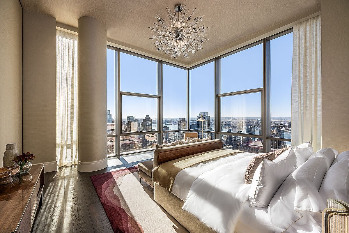 This luxurious primary bedroom features floor-to-ceiling corner windows that offer a breathtaking, panoramic view of the city skyline. The room is anchored by a plush, modern bed with neutral-toned bedding and a striking, starburst-style chandelier that serves as a focal point. A rich, patterned area rug sits atop dark hardwood floors, creating a sophisticated and serene atmosphere perfect for high-end urban living.